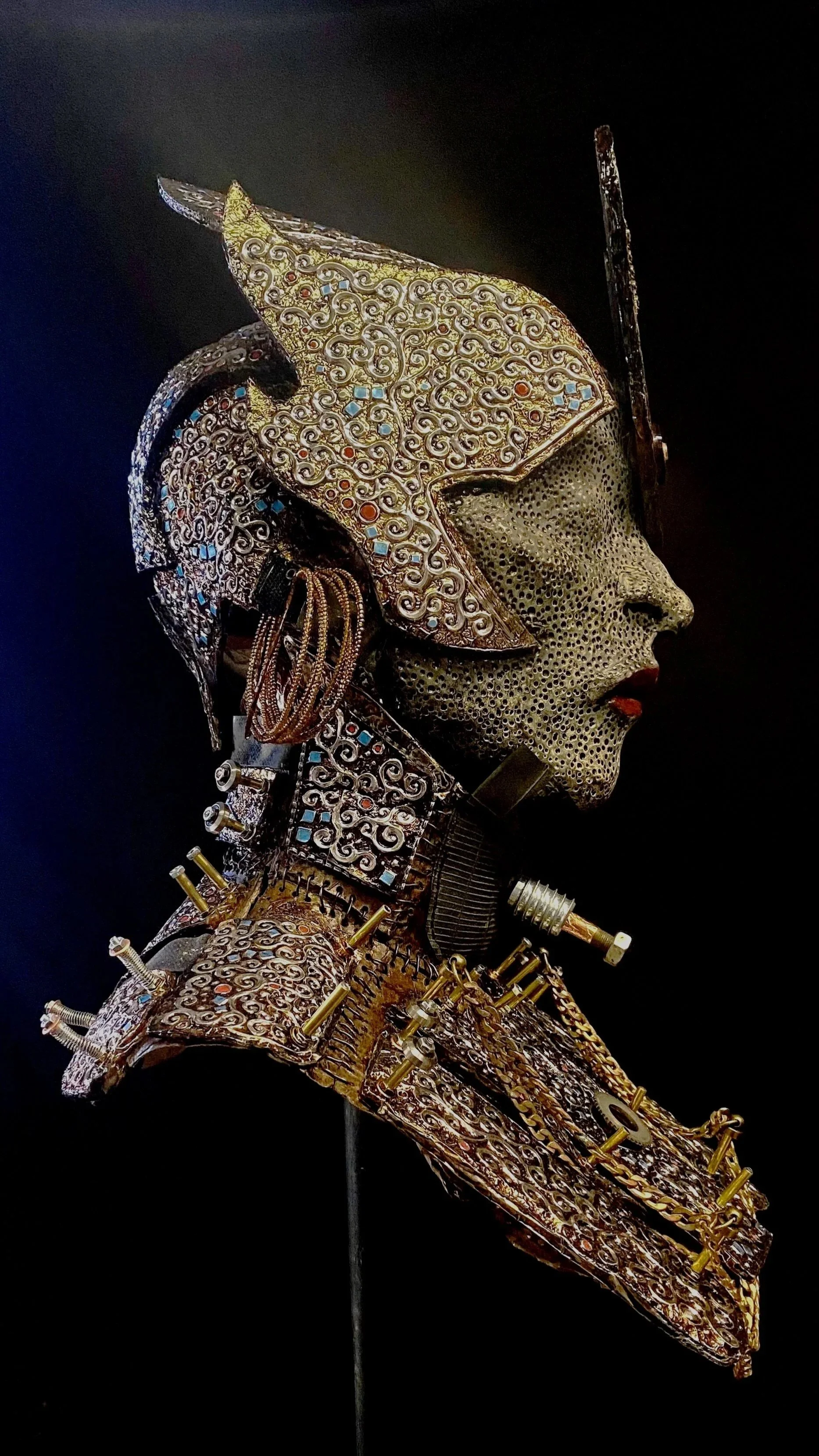 A detailed metallic sculpture of a woman’s profile with intricate patterns, blue and red accents, and decorative elements, set against a dark background.