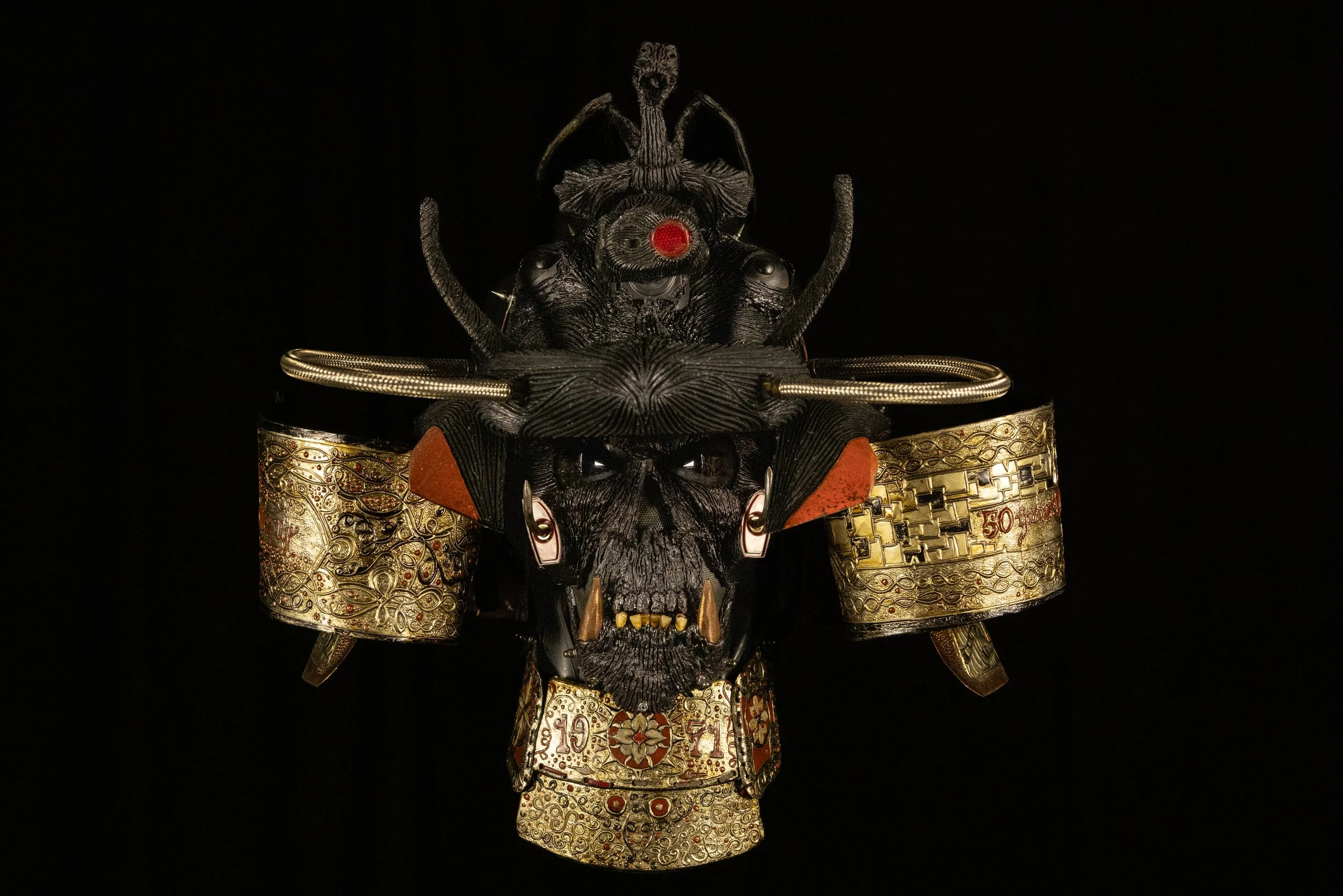 A detailed sculpture of a mythological creature with a wolf's face, adorned with gold and metallic accessories, including armor and ornaments, against a black background.