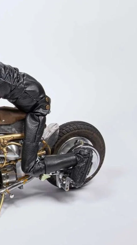 Close-up of a motorcycle with a leather seat, black leather saddlebag, and a single rear wheel, set against a plain white background.