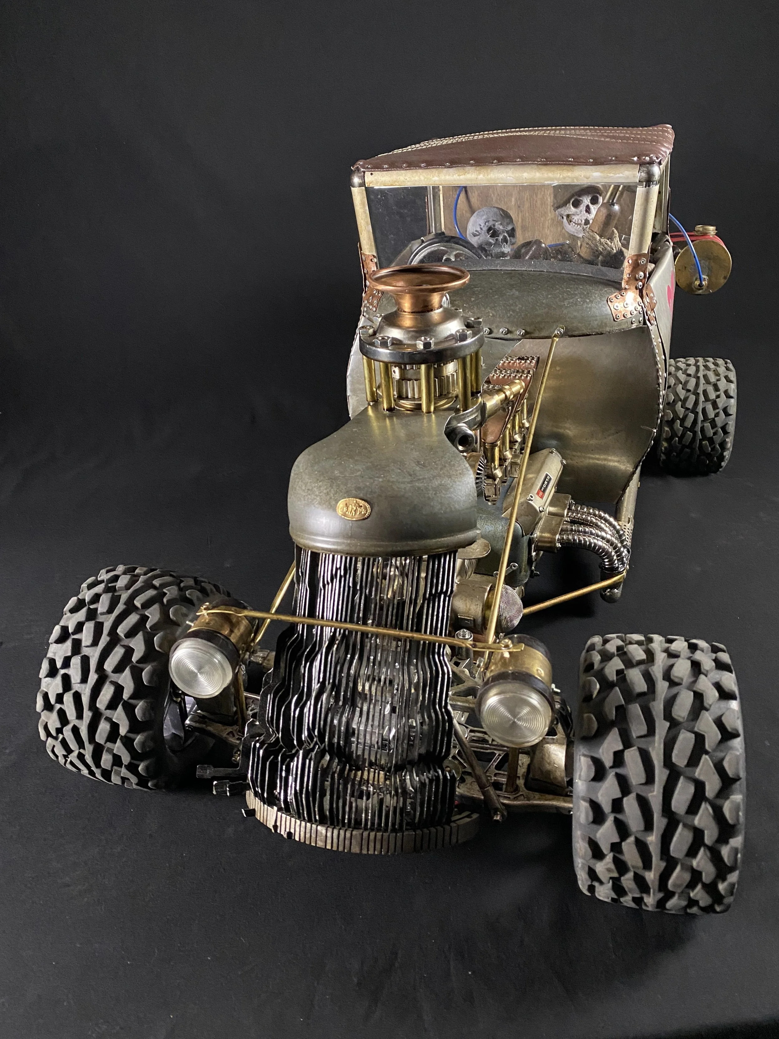 A miniature vintage hot rod car with a skull and skeleton figures in the back seat, detailed with metallic and brass elements, set against a black background.