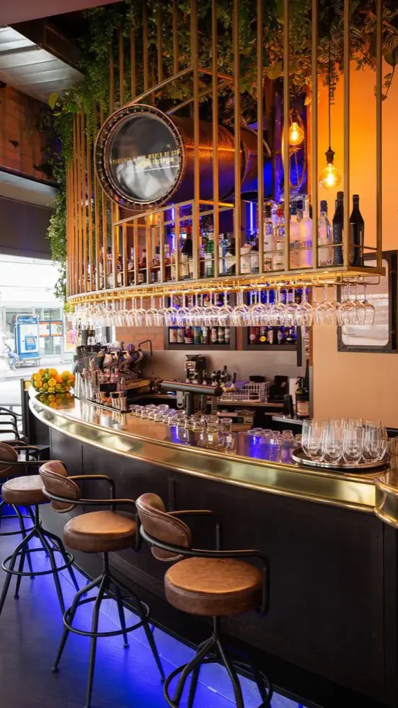 Upscale bar with a curved counter, hanging wine glasses, and a well-stocked liquor shelf, decorated with warm lighting and greenery.