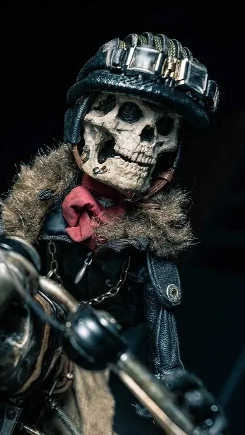 A skeleton dressed in biker gear, wearing a black helmet with metal embellishments, a leather jacket with a fur collar, and a red bandana, holding handlebars of a motorcycle.