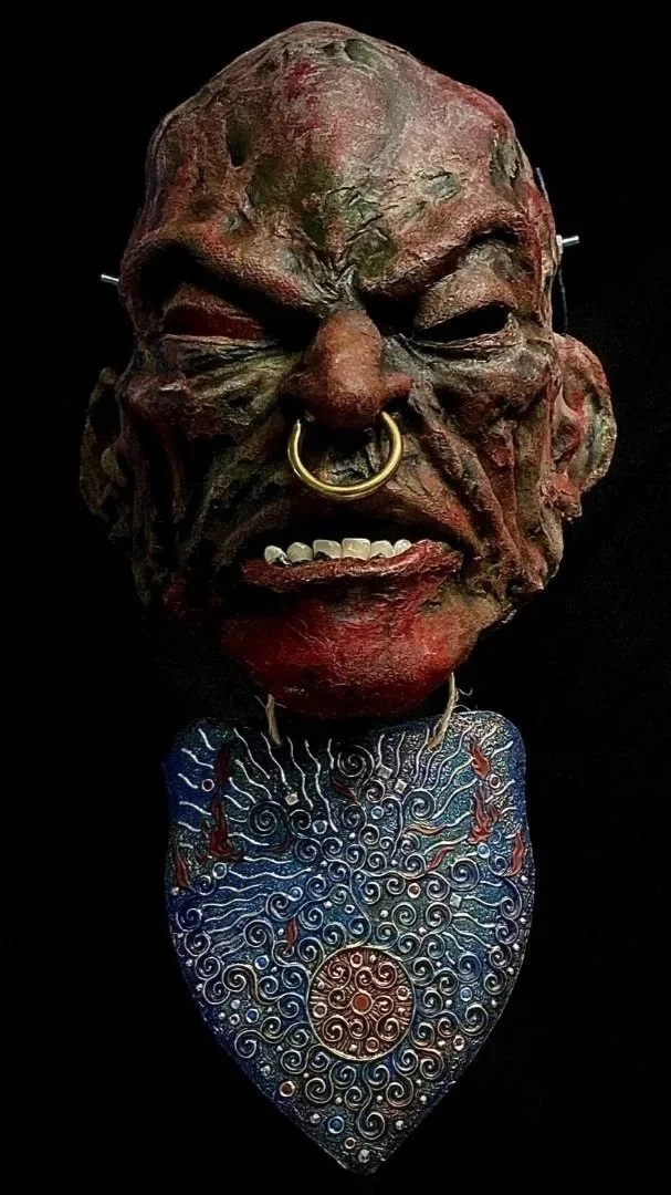 A mask or sculpture featuring a distorted, grotesque face with a large nose pierced by a gold ring, prominent teeth, and textured skin. The face is mounted on a decorative, swirling pendant with intricate patterns and blue, red, and gold accents.