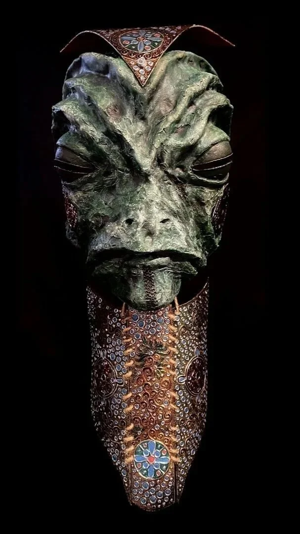 A tribal mask with a textured, greenish face featuring prominent brow ridges and closed eyes, decorated with colorful, intricate patterns and adorned with beads and circular motifs.