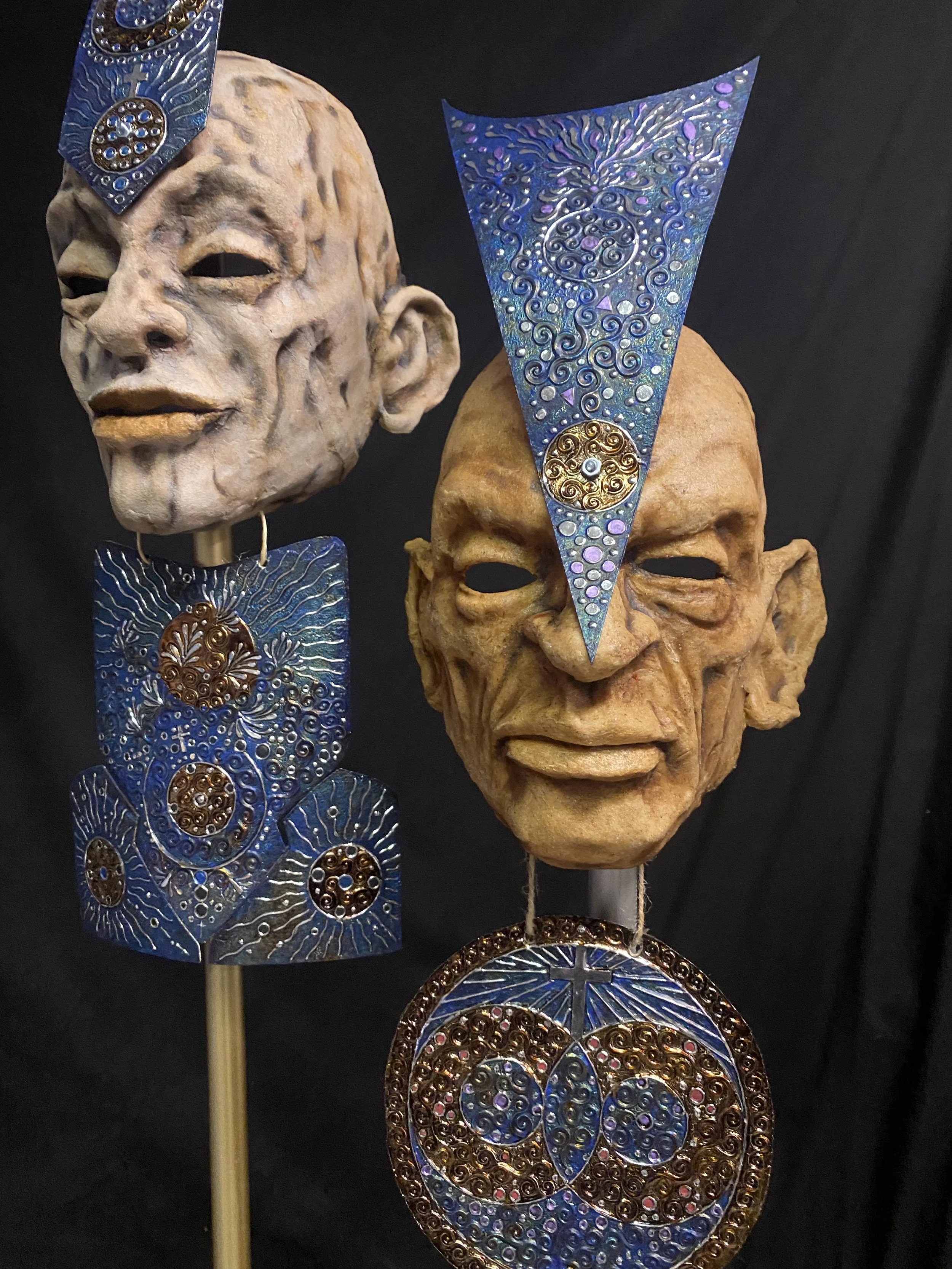 Two tribal masks with large facial features and elaborate blue and gold headdresses or adornments, mounted on sticks against a dark background.