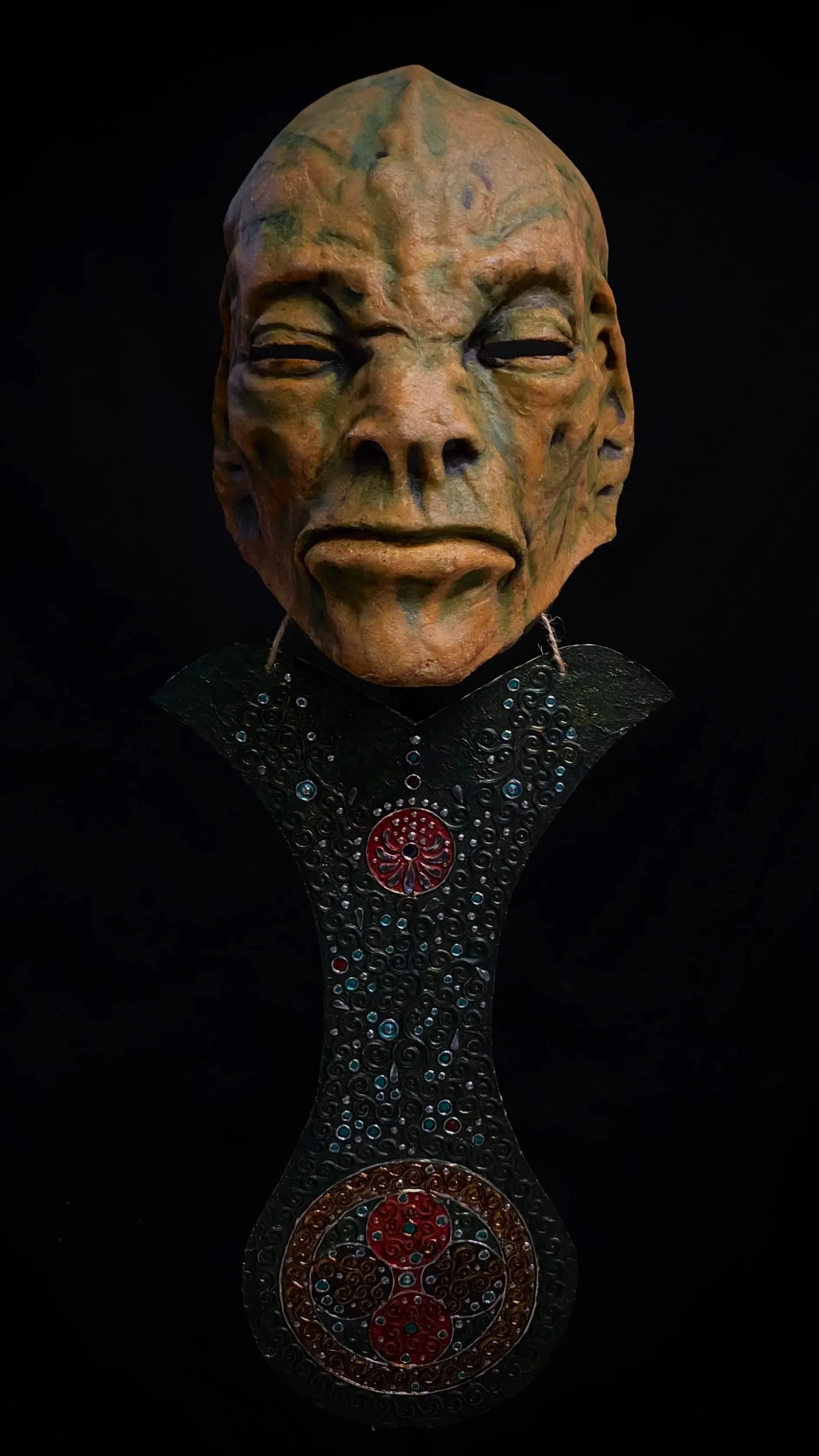 An artistic sculpture of an elderly man's face with detailed features, attached to a decorated black necklace or collar against a black background.