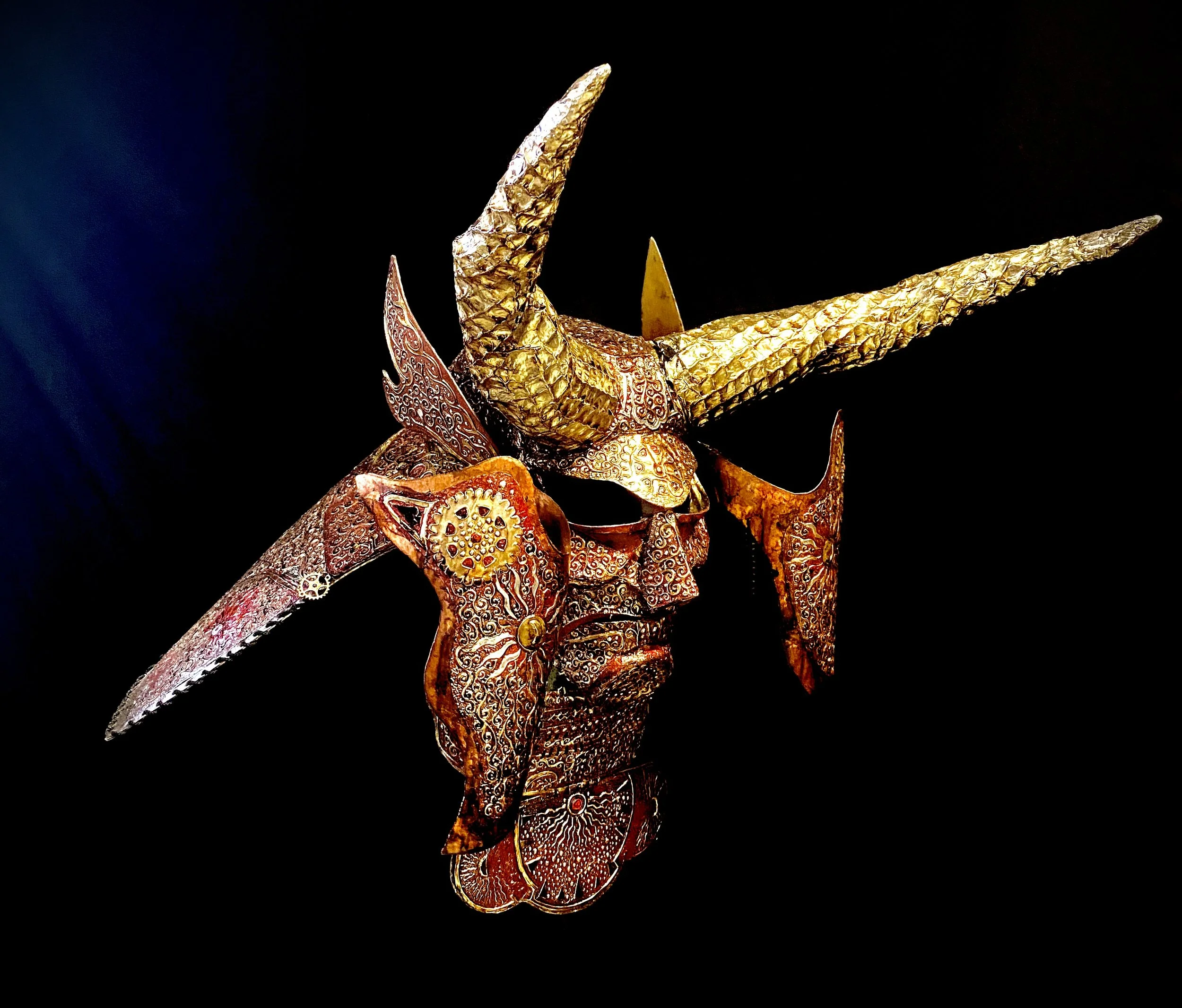 Intricately designed art sculpture of a warrior mask with elaborate metal embellishments and  large golden horns extending from the forehead, set against a black background.