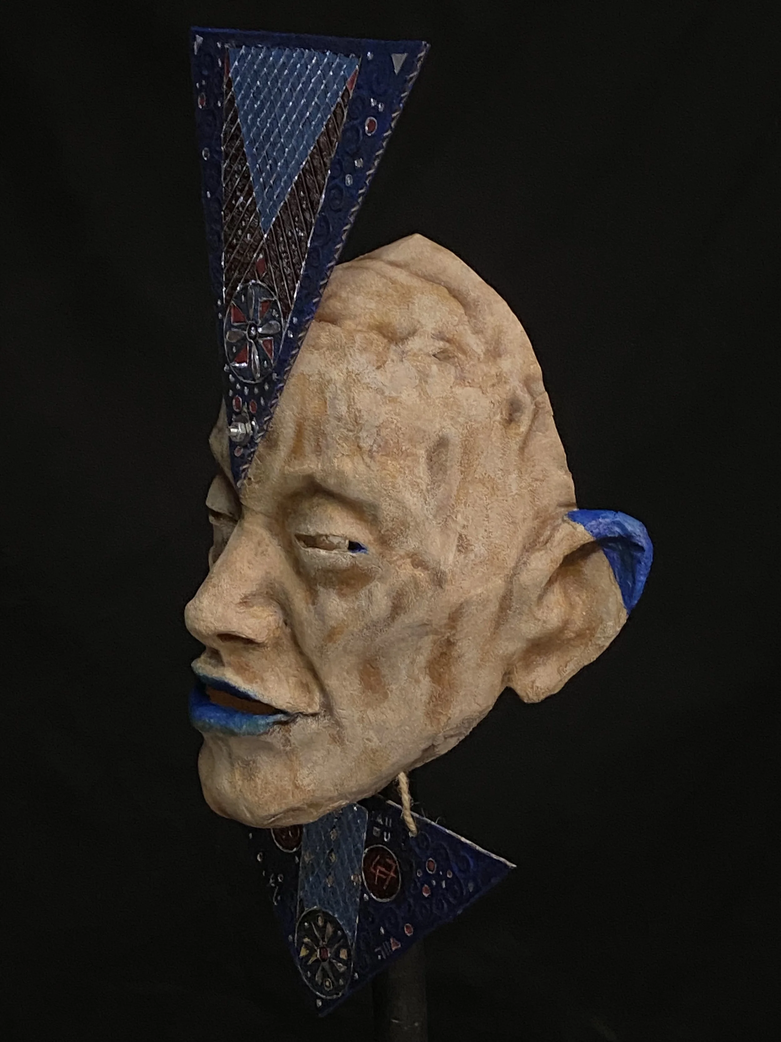 A mixed media sculpture of a human face with exaggerated features. The face has blue lips, slightly squinted eyes, and prominent cheekbones. The sculpture combines a realistic textured face with abstract blue and patterned accessoires, including a ta