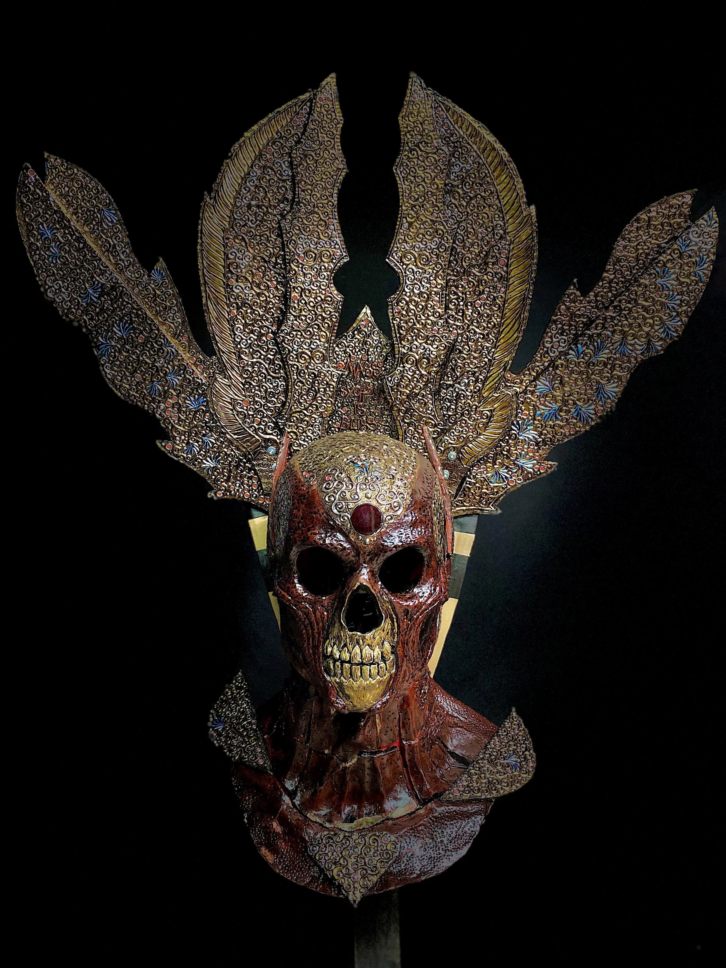 A decorative art piece featuring a skull with an ornate headdress or crown made of patterned gold and multicolored metalwork, set against a black background.