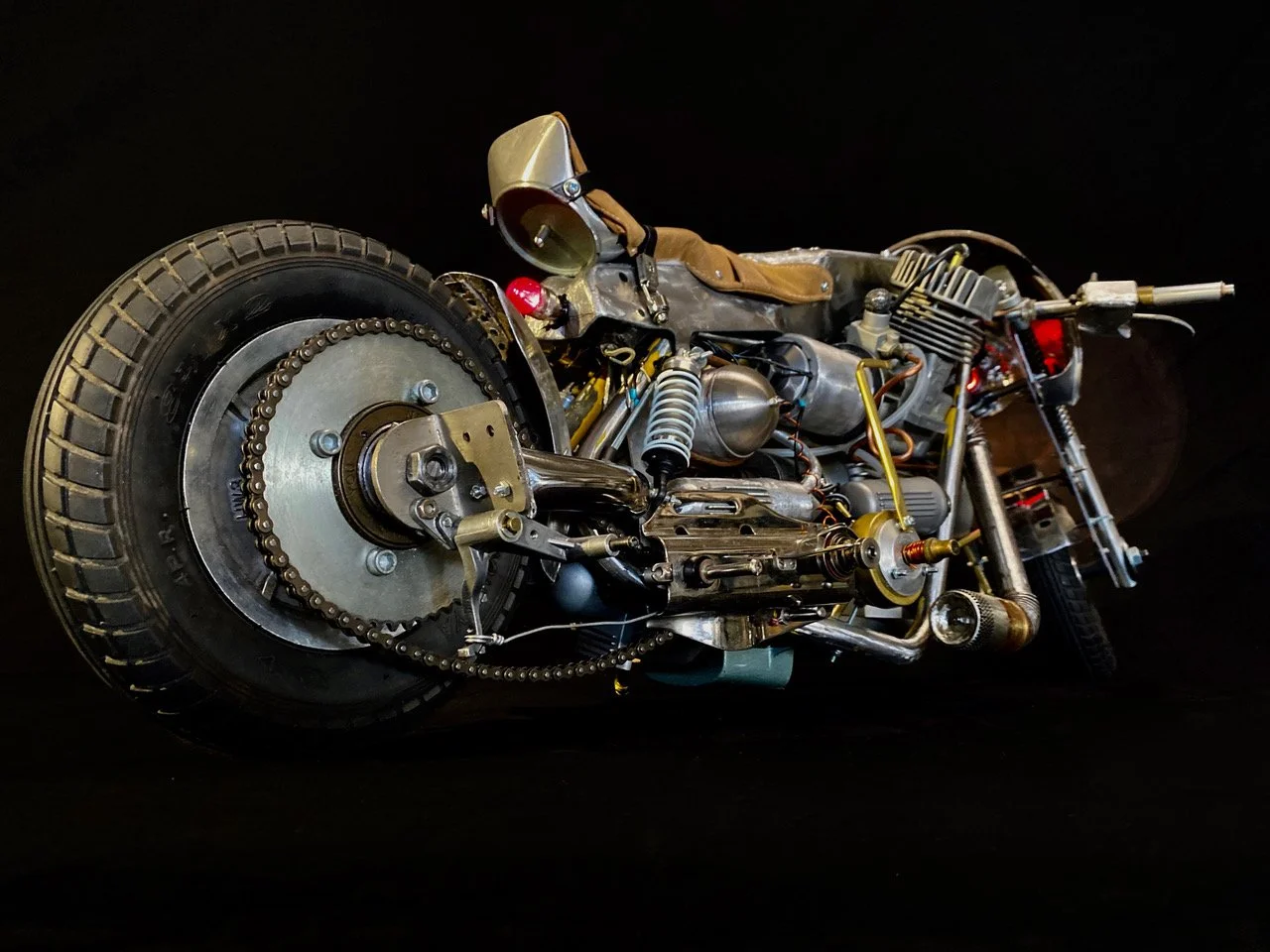 A detailed, vintage-style motorcycle frame and engine with exposed metal components, a prominent chain drive, and a tan leather seat, set against a black background.