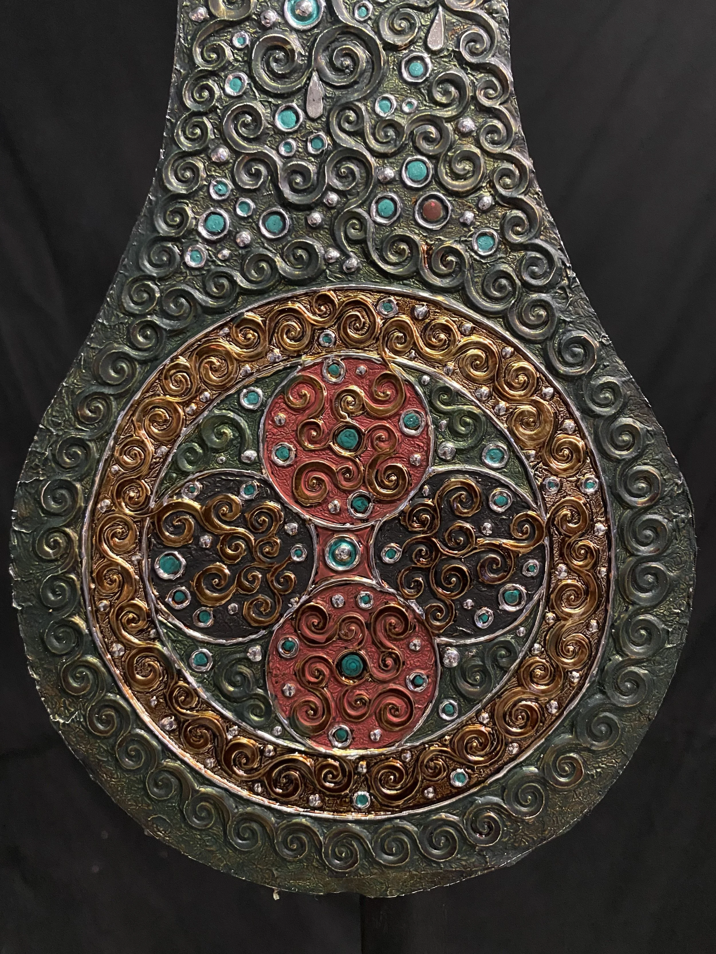 A decorative ornament with intricate swirling patterns in gold, silver, and red, adorned with small turquoise and white stones.