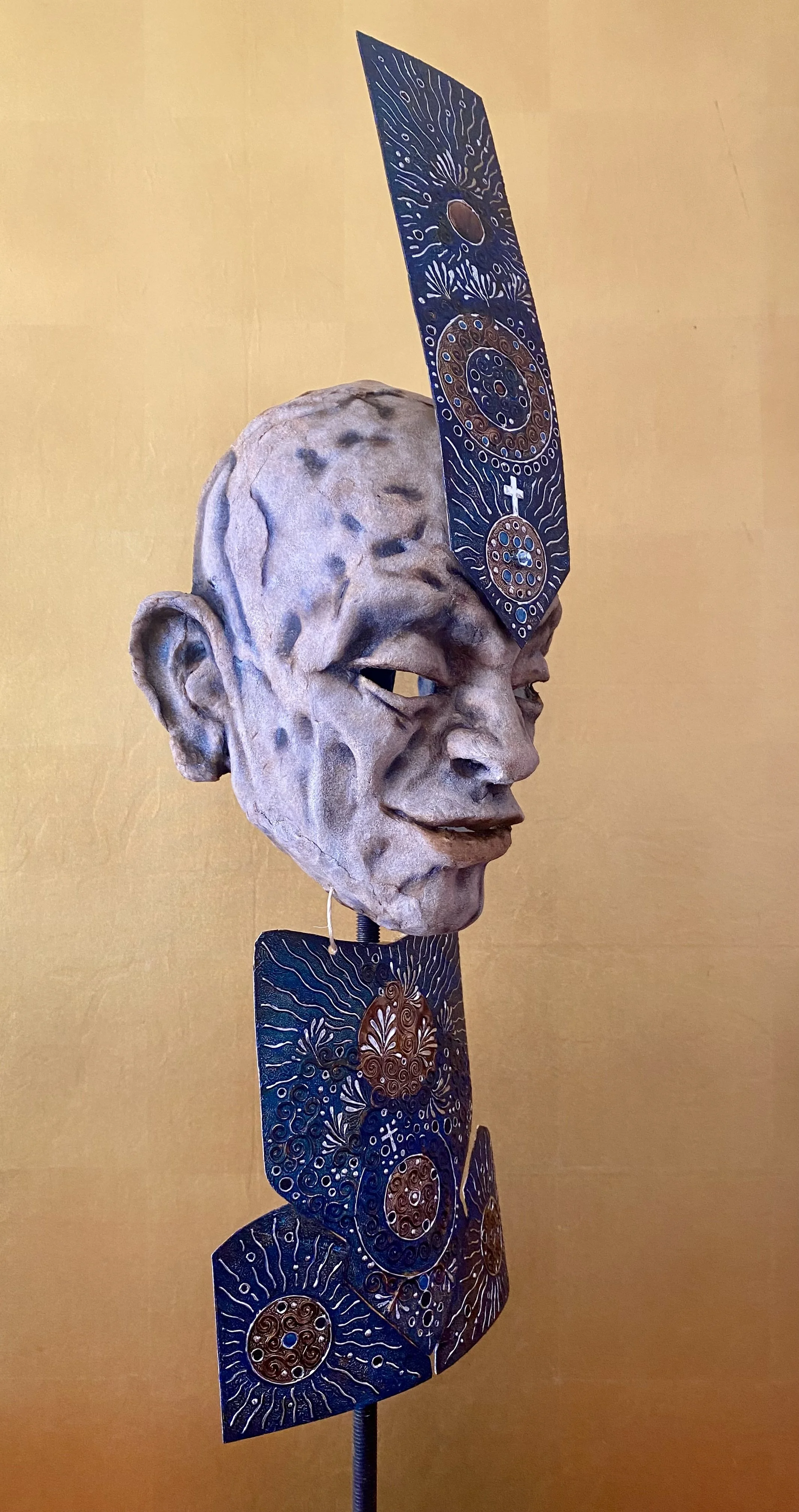 A sculpture of a human face with a textured, clay-like appearance, mounted on a stand. Two rectangular, decorated panels with intricate designs and patterns, primarily in blue, brown, and white, extend vertically from the sculpture's head.