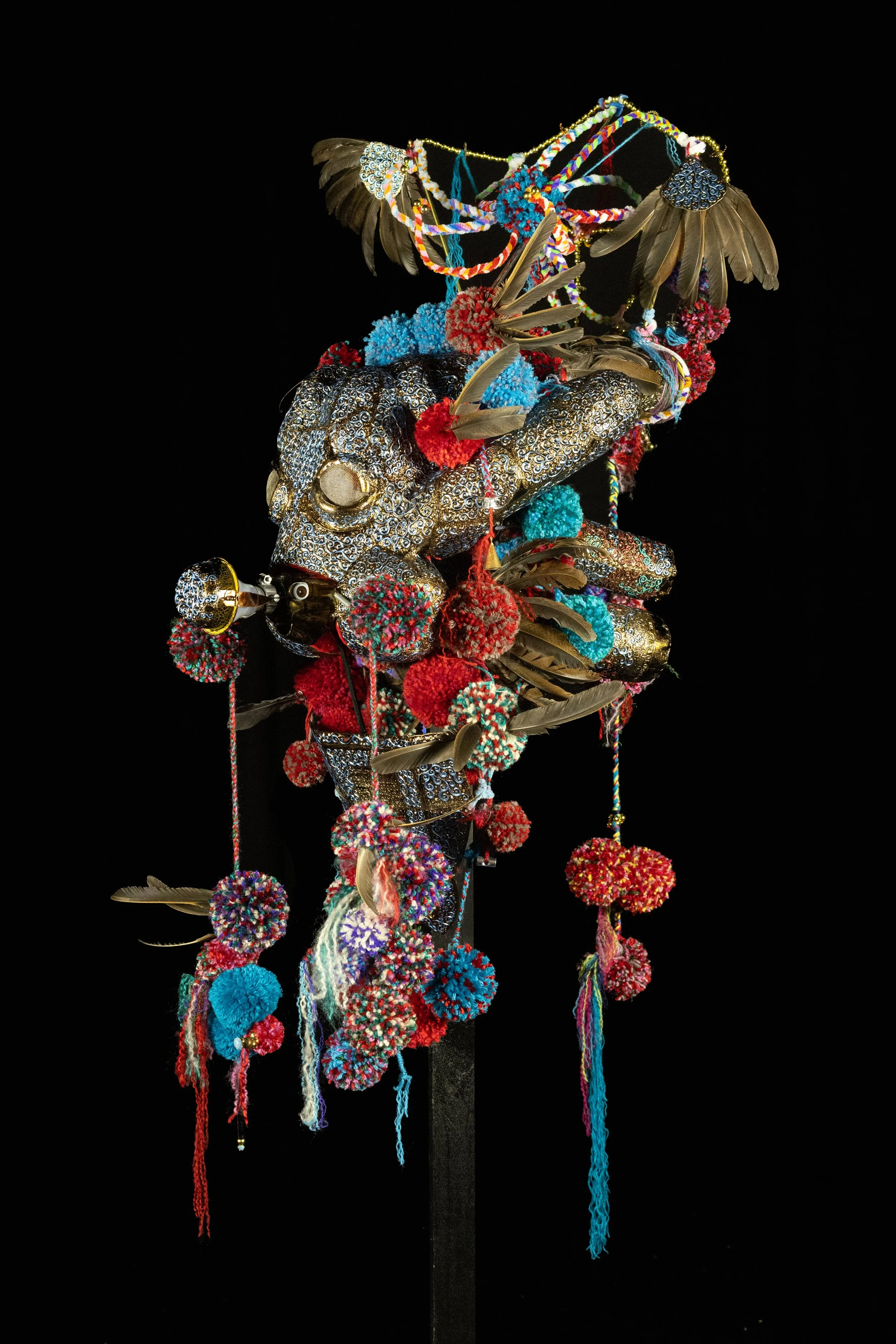 An art installation of a shaman mask composed of various objects including feathers, colorful yarn, and metallic elements against a black background.