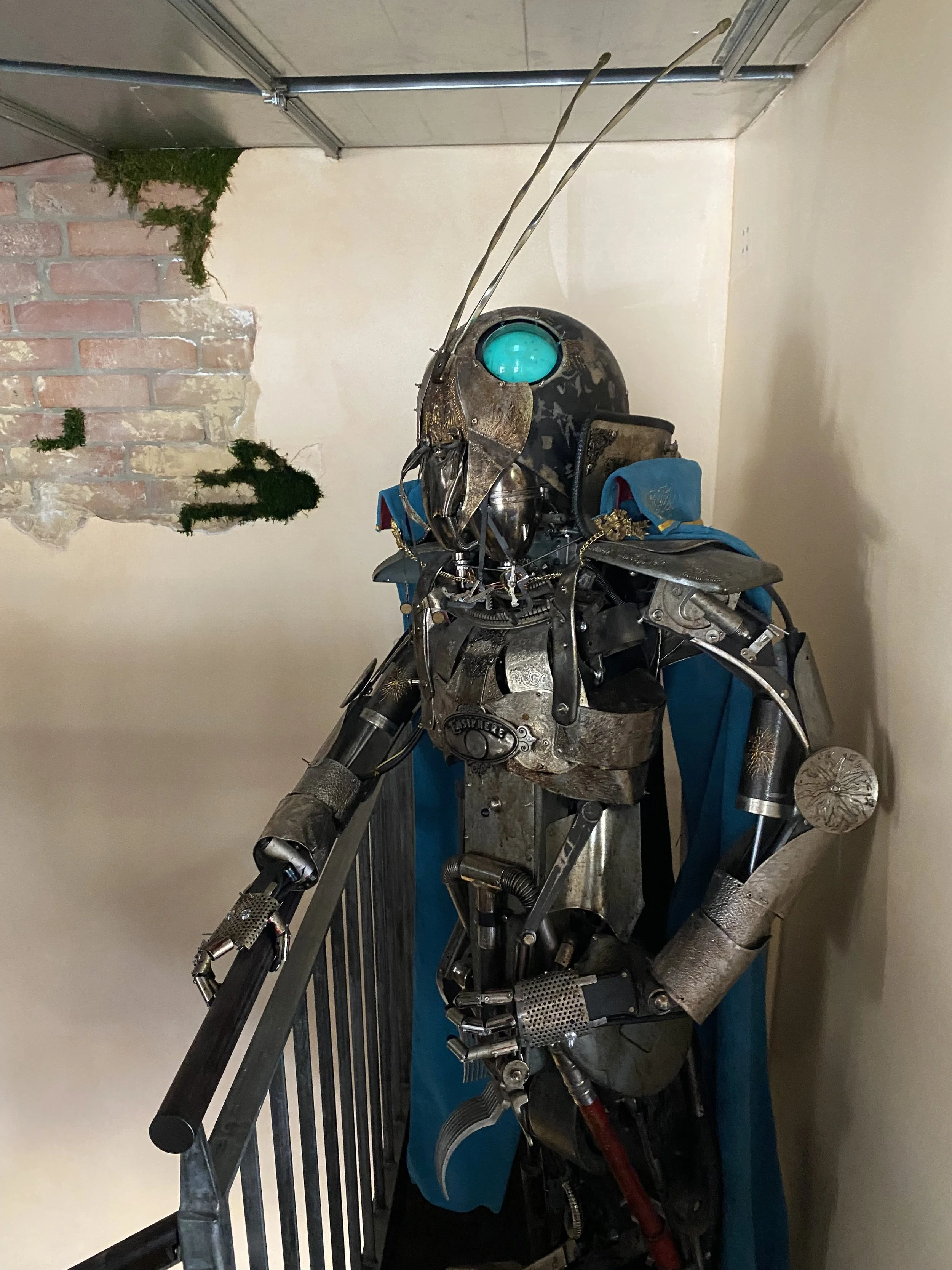 A humanoid robot with metallic parts, a glowing blue eye, and antennae, wearing a blue cape, displayed indoors against cream-colored and brick wall backgrounds.