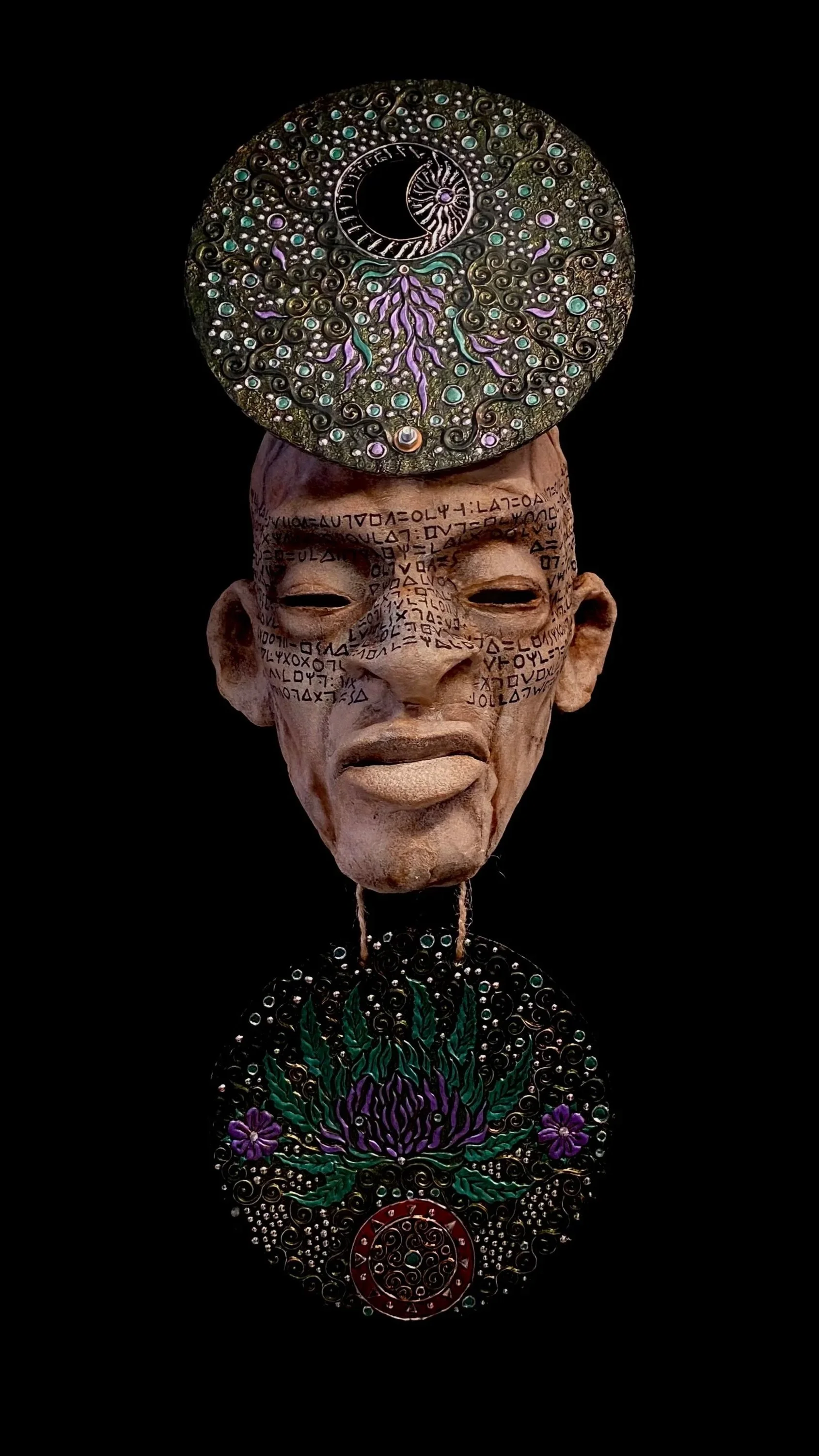 A tribal-style art piece featuring a carved face with mysterious markings, topped with a circular, decorated disc with celestial and floral motifs, connected by a string to a similar decorated disc at the bottom.