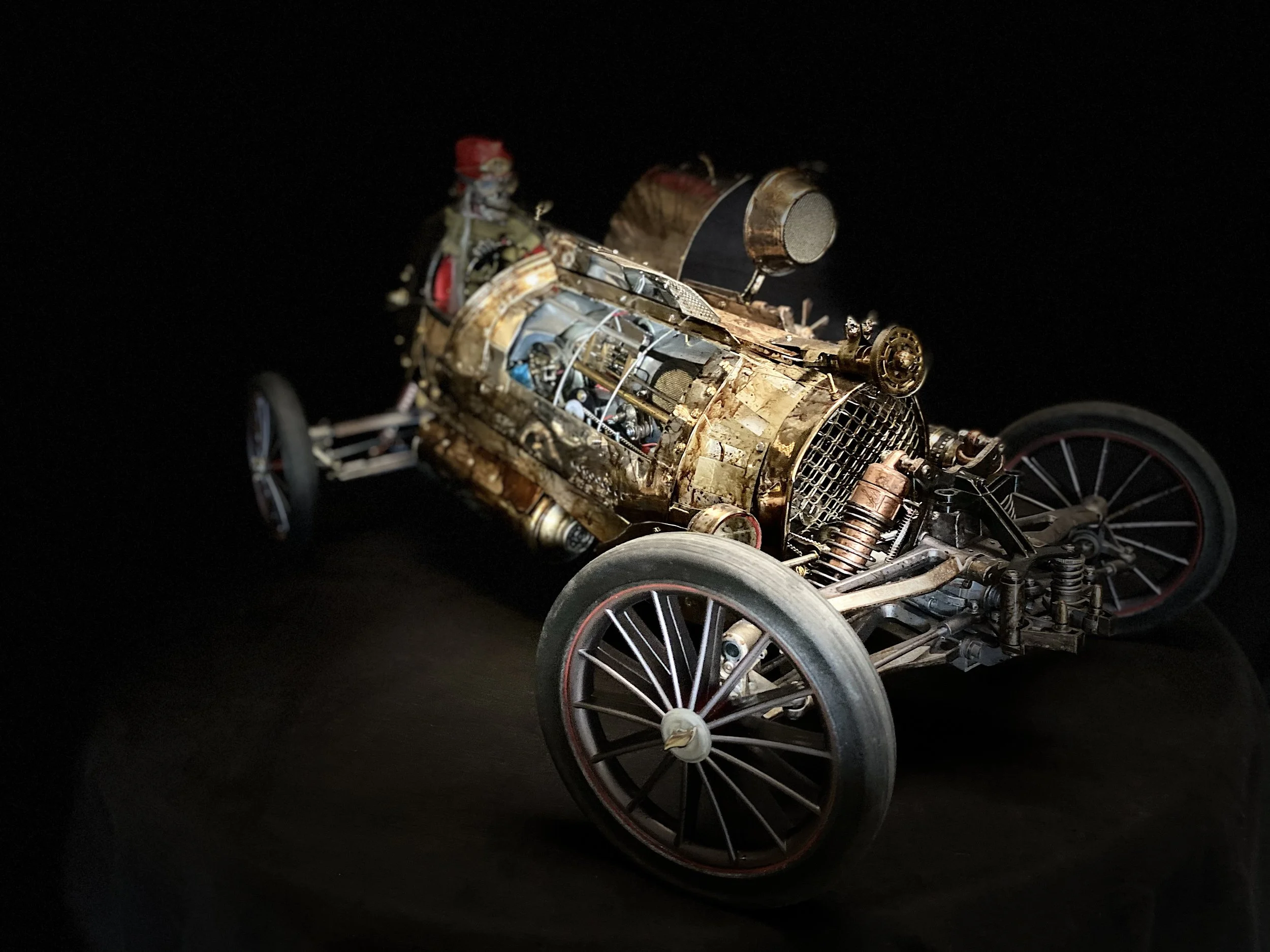 A detailed model of a vintage race car with exposed mechanical parts and a small driver figure in a racing suit and helmet inside the cockpit, set against a black background.