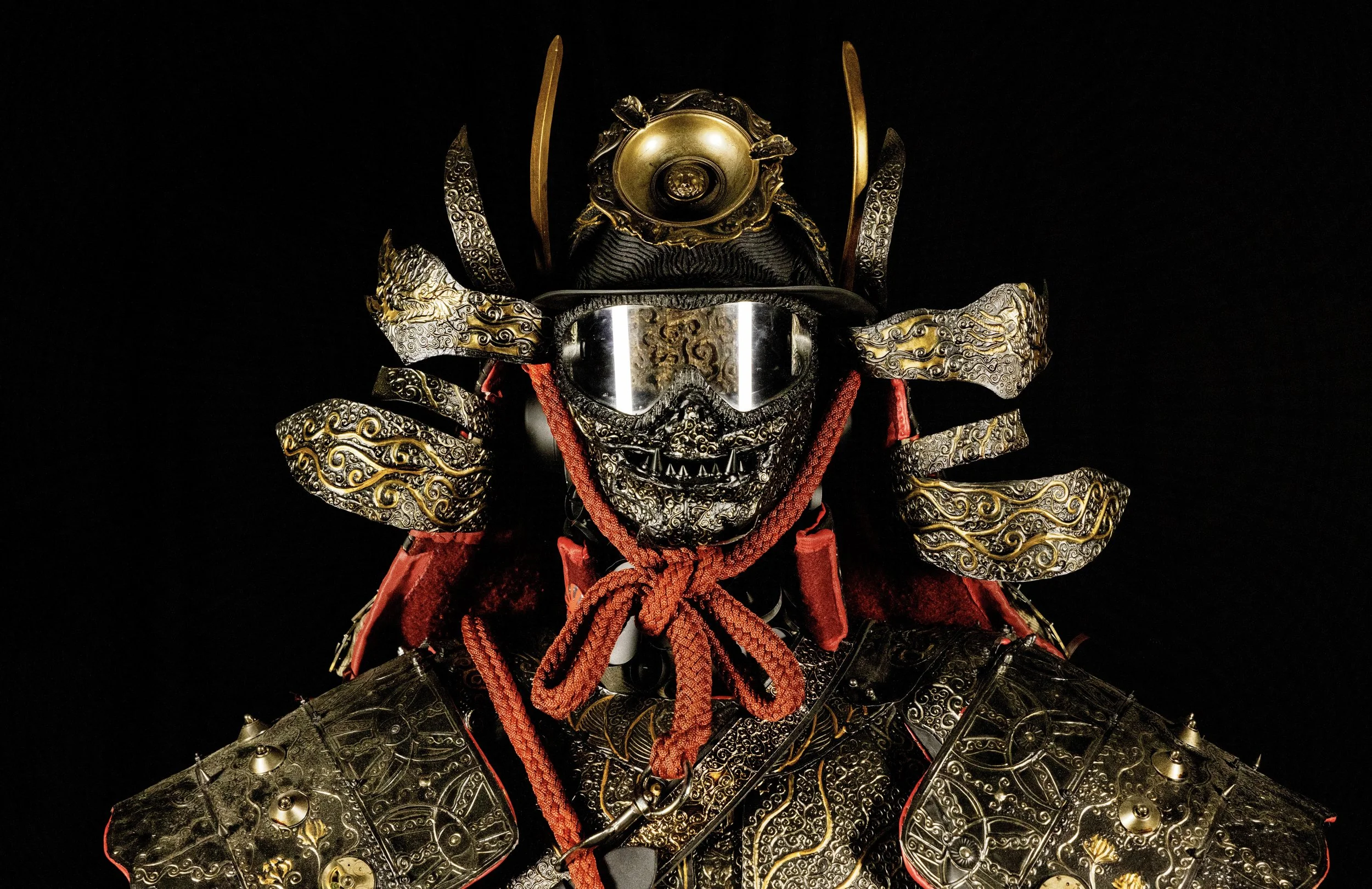 A detailed suit of armor with ornate gold and black engravings, a red cloth tied around the neck, a metallic mask with goggles, and an elaborate helmet with gold accents and horns.
