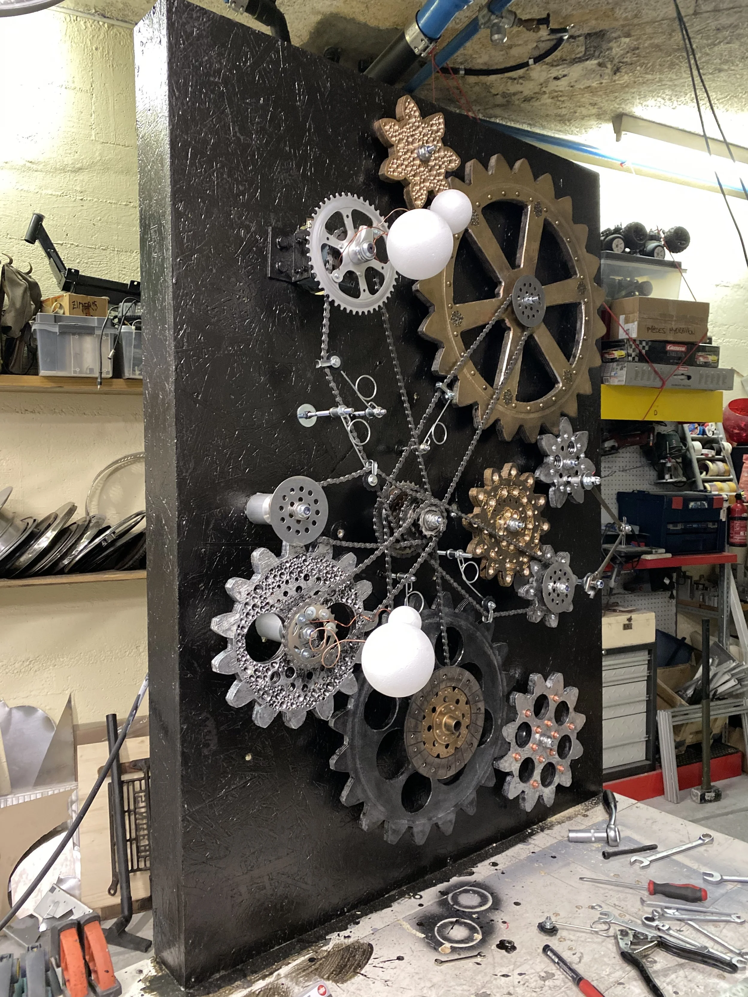 A gear wall art piece made of metal gears, chains, and decorative objects on a black board in a workshop.