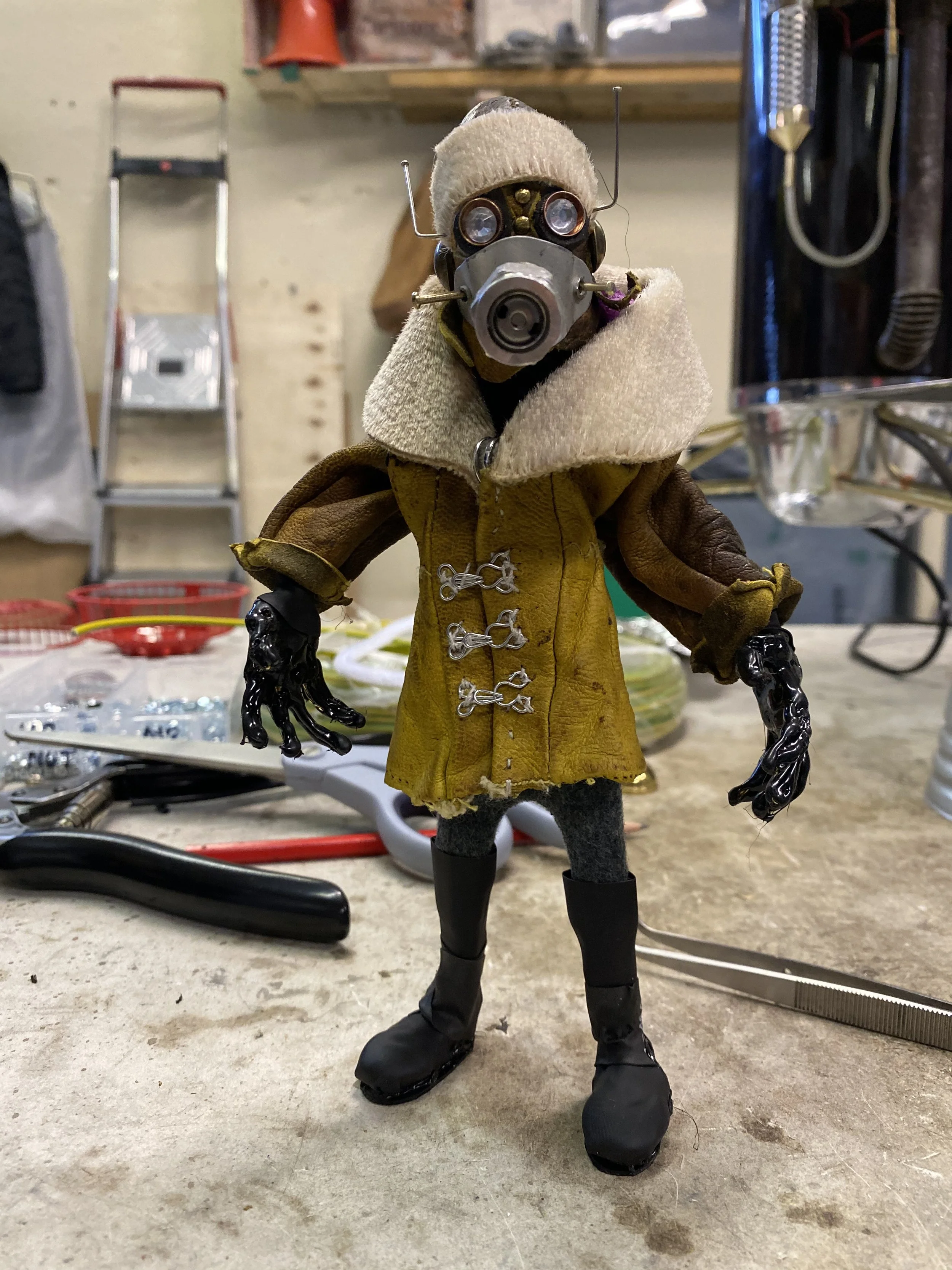 A humanoid robot figure dressed in a yellow coat with metal clasps and black boots, wearing a beige hat and a face mask, stands on a workbench in a workshop.