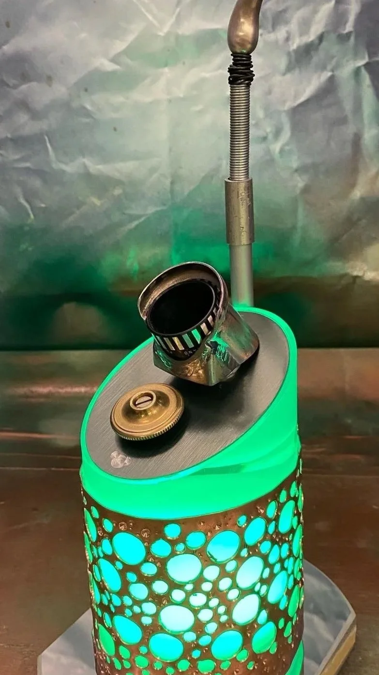 A close-up of a decorative lamp with a metallic attachment on top, a gold-colored screw cap, and a metallic cylinder. The lamp has a perforated metal cover with glowing green light shining through circular holes in the cover.