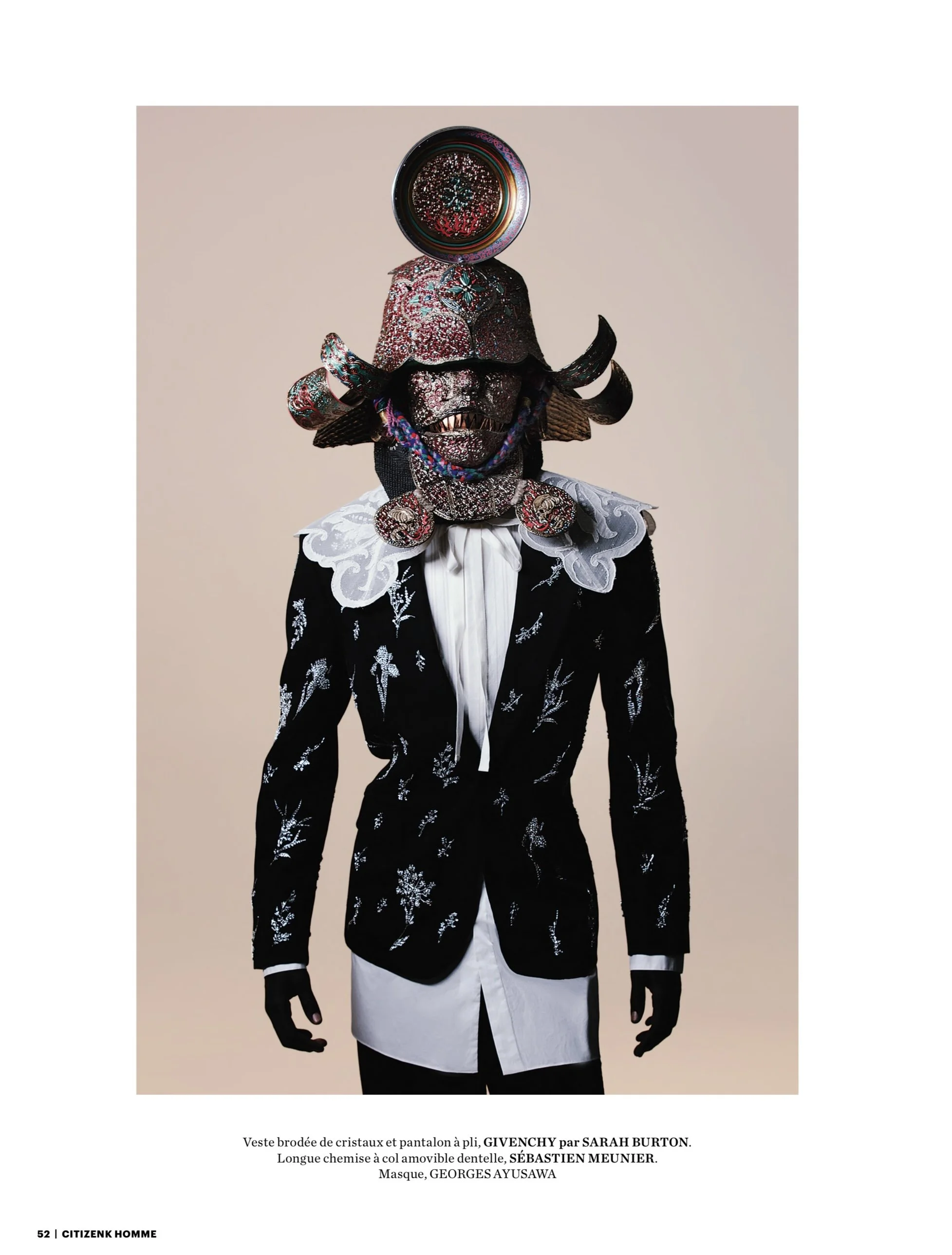 A fashion model wearing an elaborate, artistic costume with a large, ornate mask adorned with crystals and metallic elements, a crystal-embellished jacket, and a white shirt with lace collar detailing.