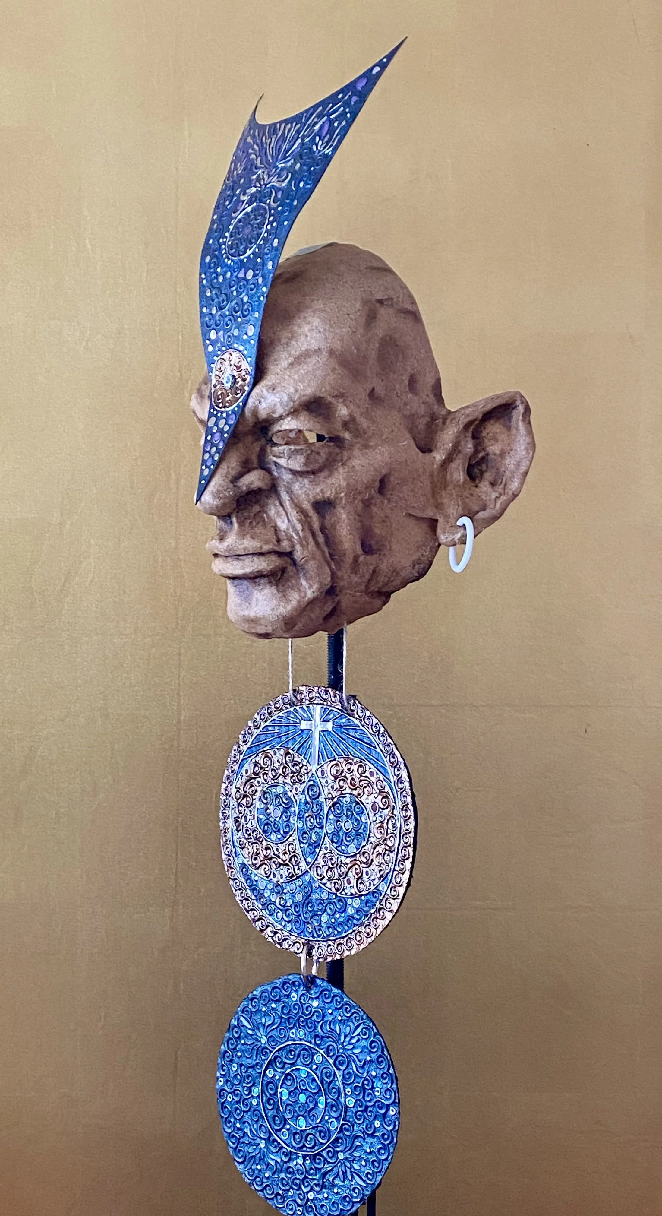 A sculpture of a human head with large ear, earring, and a blue abstract hat, attached to a stick with circular blue and brown patterned ornaments hanging below.