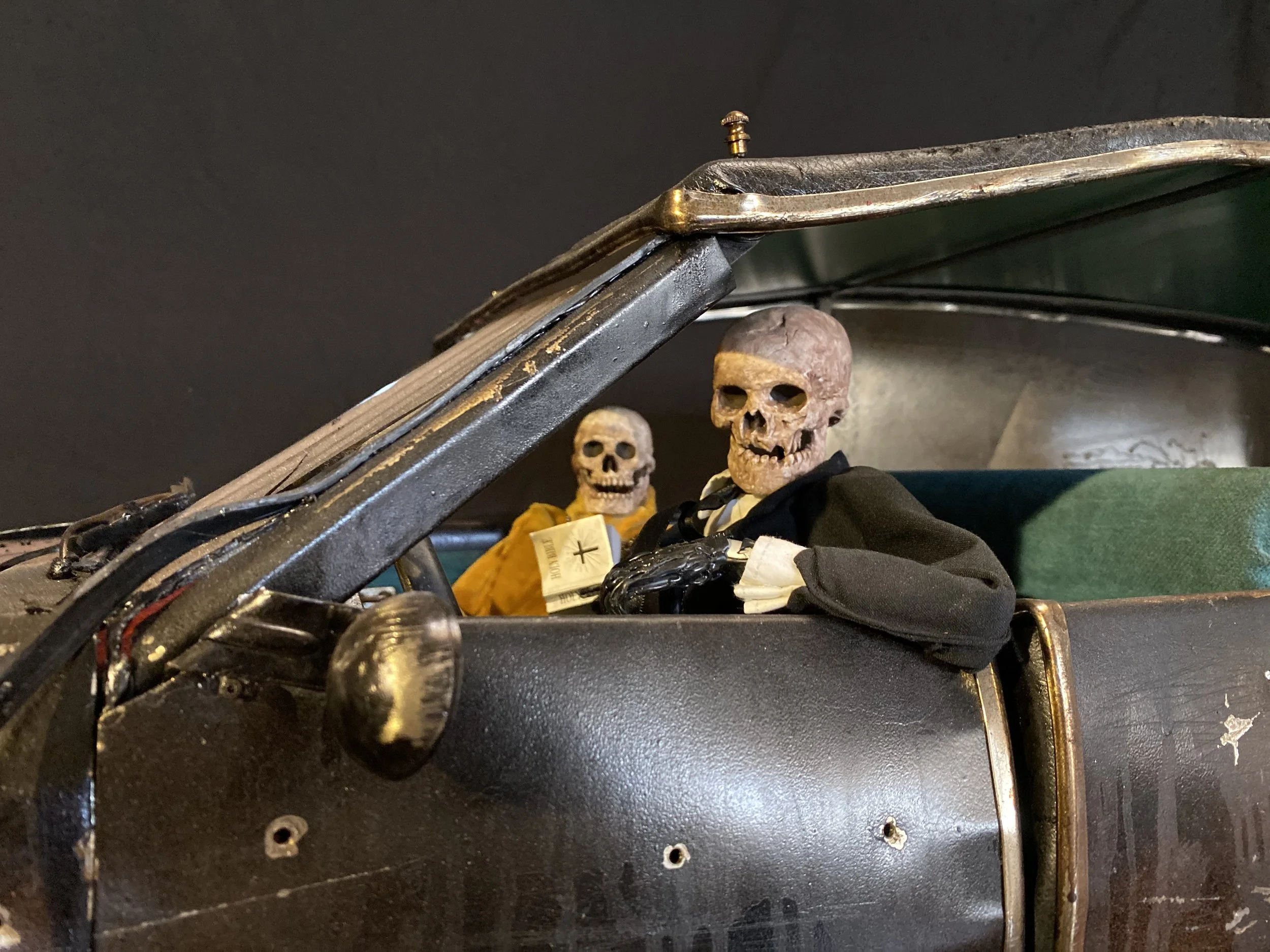 Two skeleton figures dressed in black suits sitting inside a vintage black car.