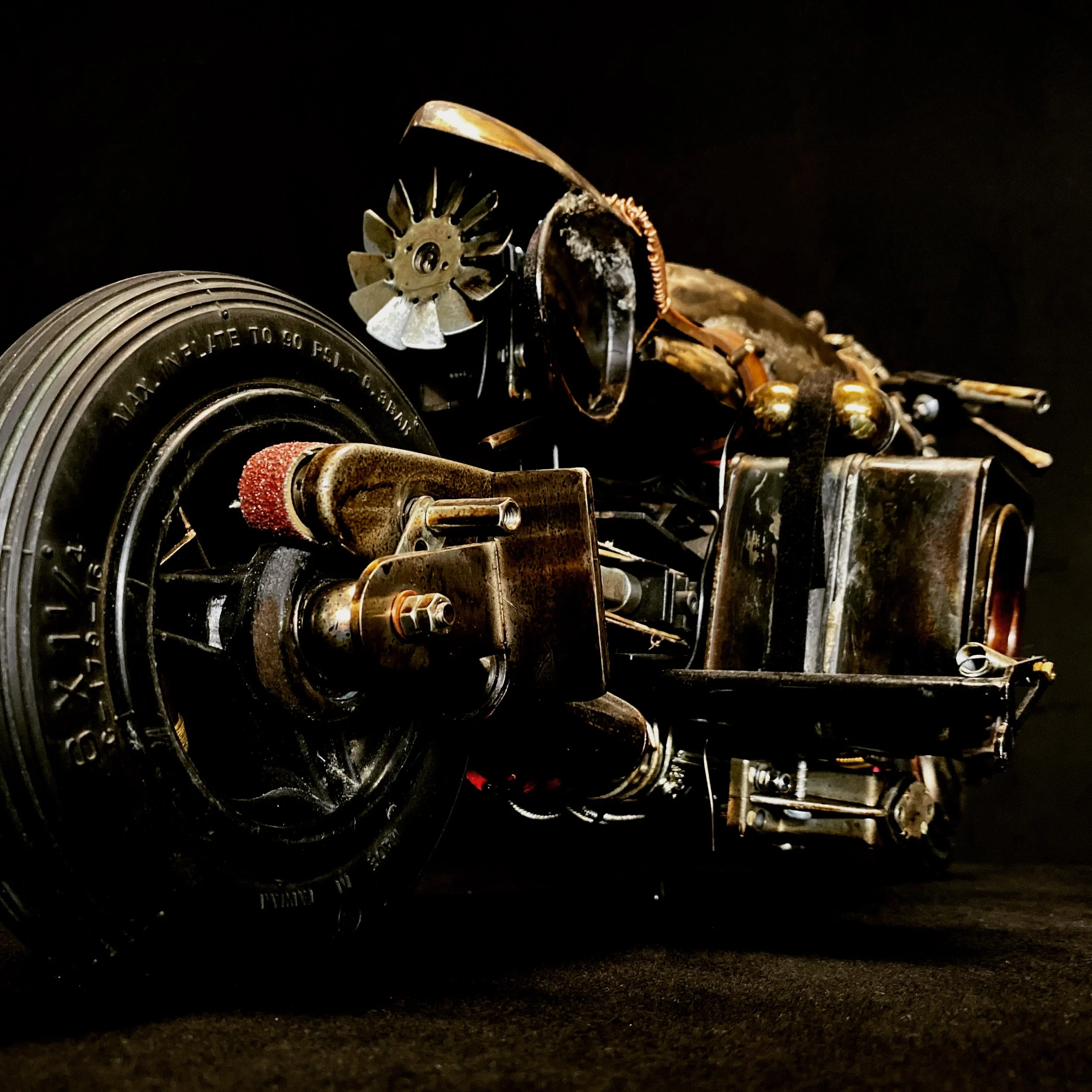 Metal robotic vehicle or sculpture with gears, engine parts, and tire, set against a black background.
