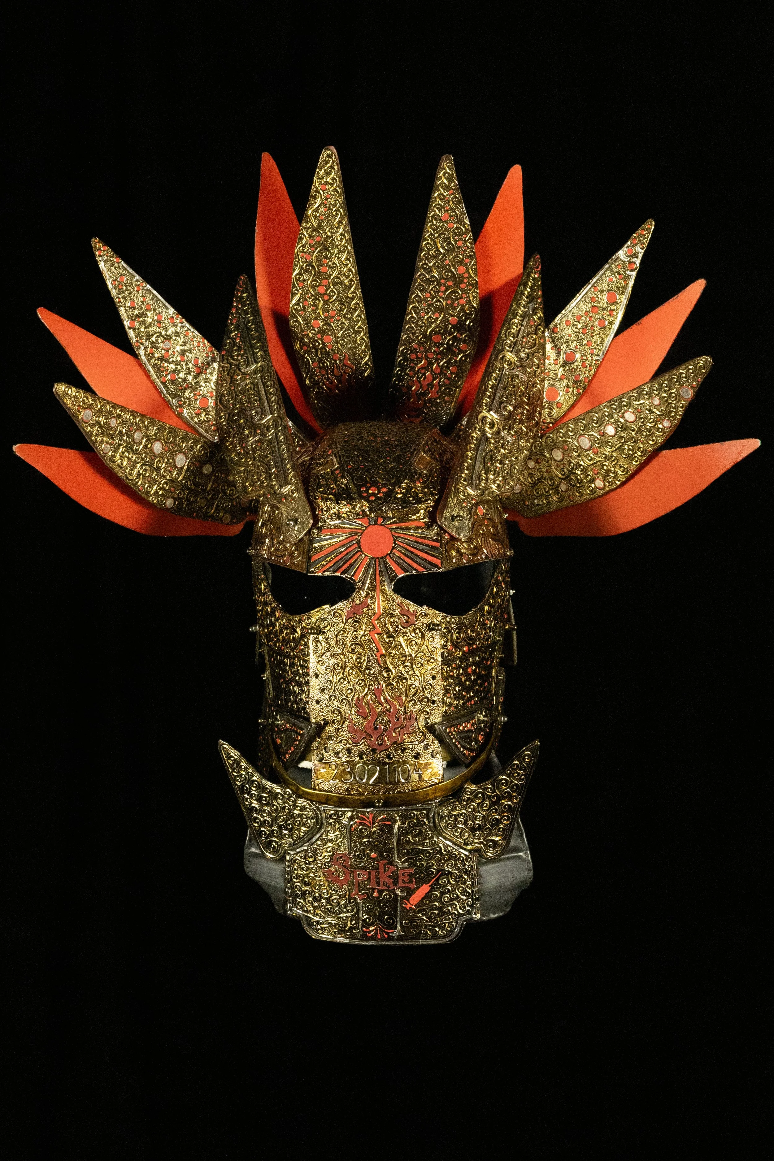 A decorative skull mask with intricate gold detailing, orange and gold feather-like structures, and red accents, displayed against a black background.