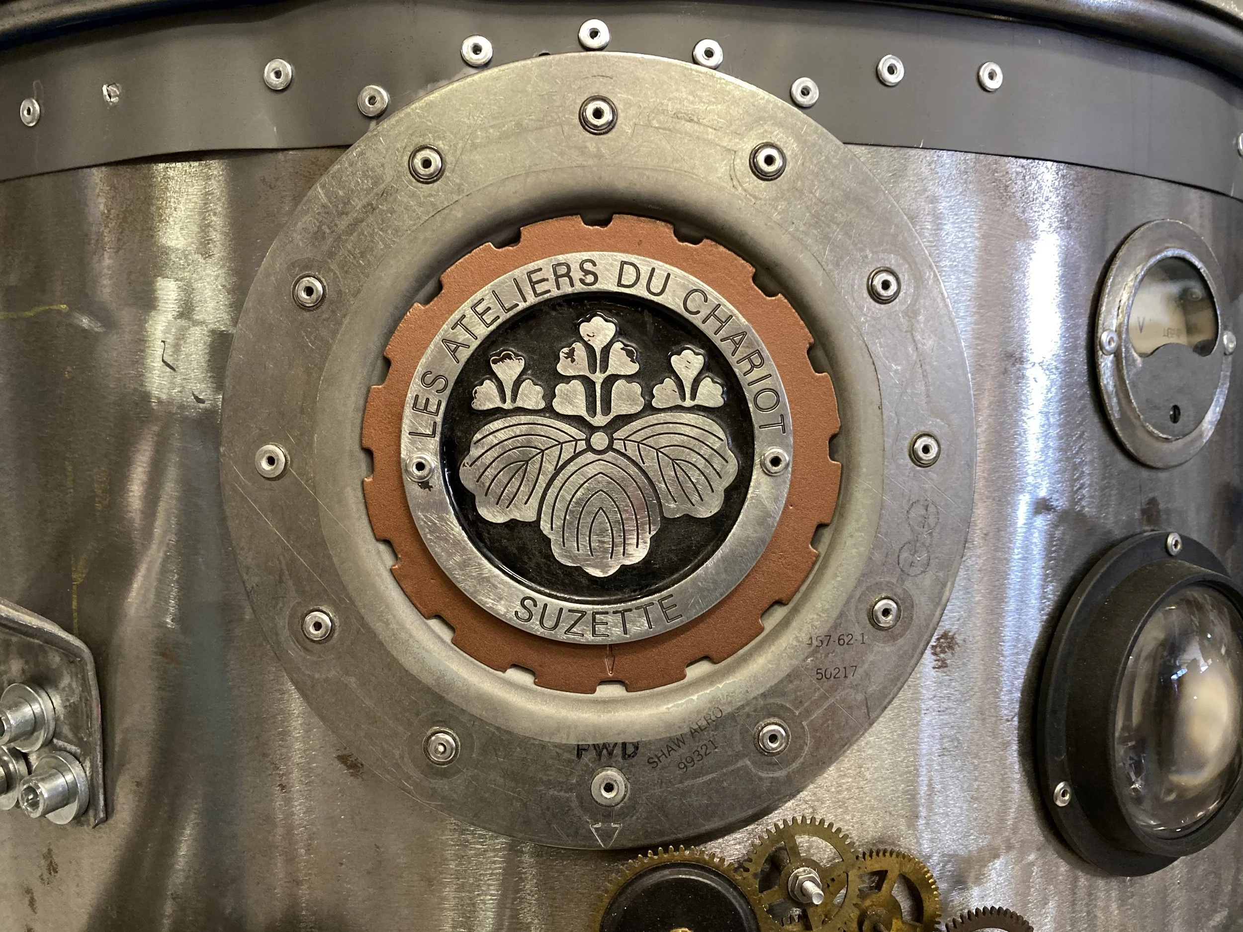 Close-up of a metal machine part with a central emblem that reads 'LES ATELIERS DU CHARIOT SUZETTE' and features a butterfly design.