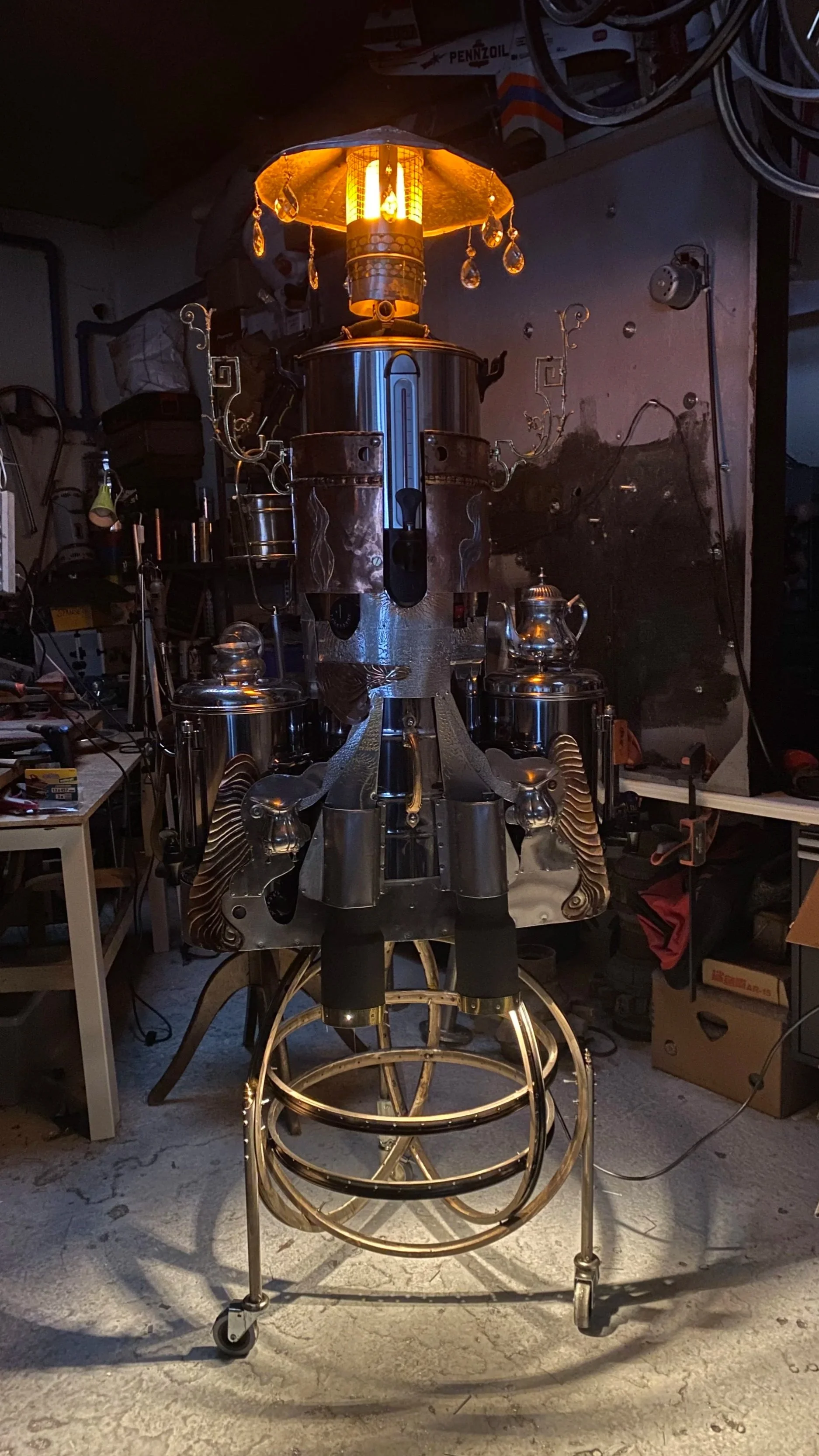 A steampunk-style lamp made from metallic parts placed on a workbench in a cluttered garage or workshop. The lamp has a filament bulb with dangling crystal decorations and is supported by a decorative metal frame with wheels.