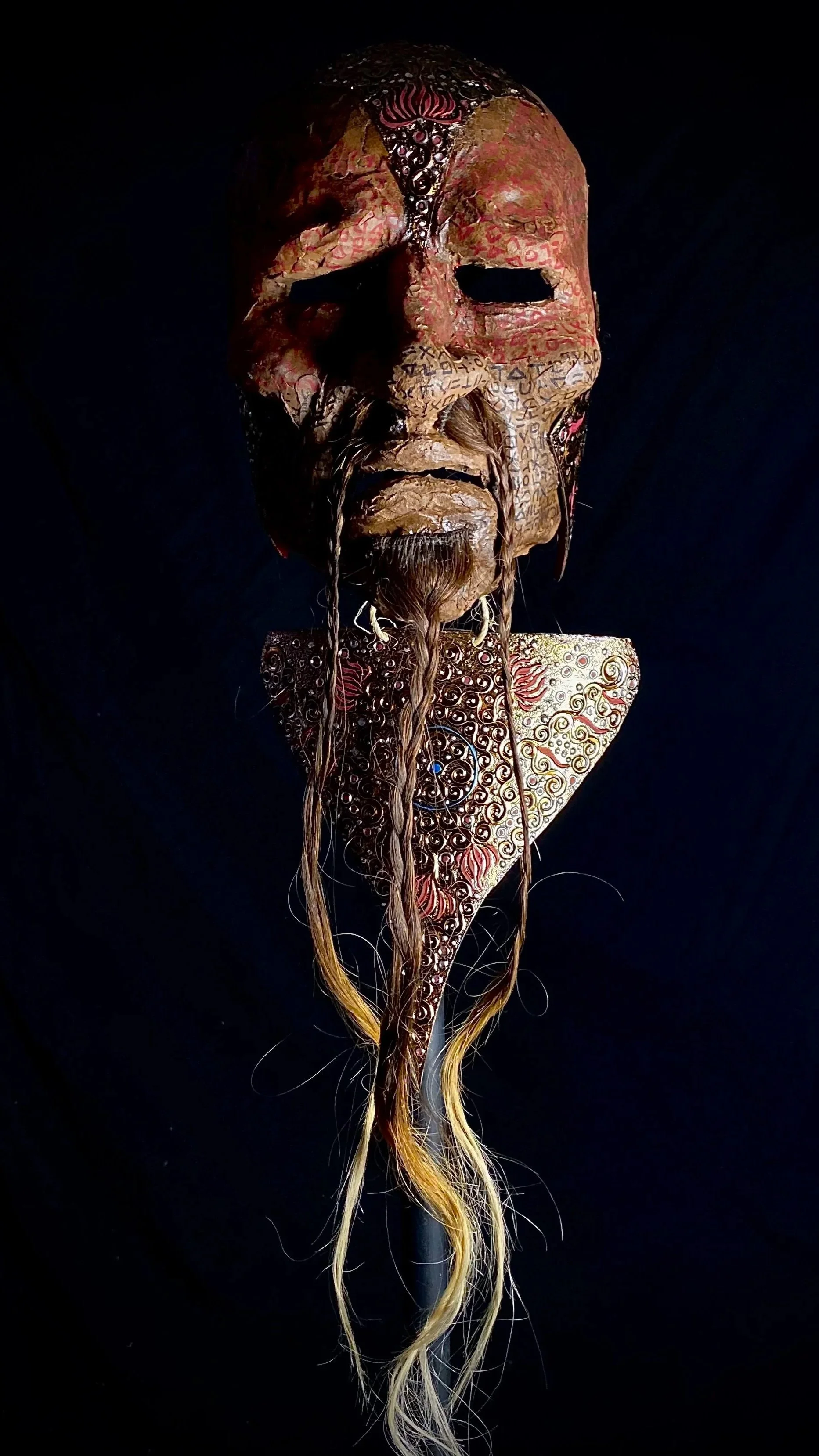 A traditional carved wooden mask with intricate designs, long gray and yellow hair, and facial features with a mustache and beard, set against a dark background.