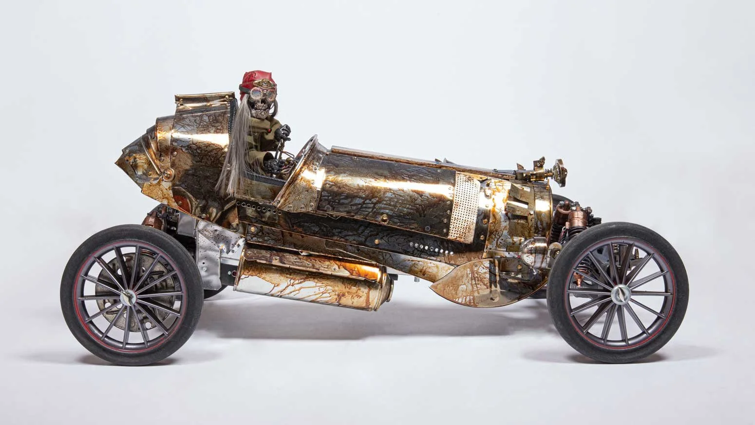 A detailed steampunk-style model of a vintage race car with metallic and rusted finishes, featuring large spoked wheels and a skeleton figure dressed as a race car driver in the driver’s seat.