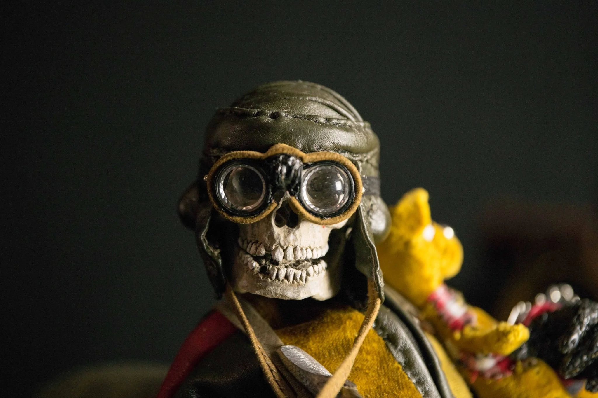 A skeleton wearing a leather pilot helmet and goggles, with a yellow shirt and scarf. The skeleton is riding a motorcycle with a teddy bear in the background.