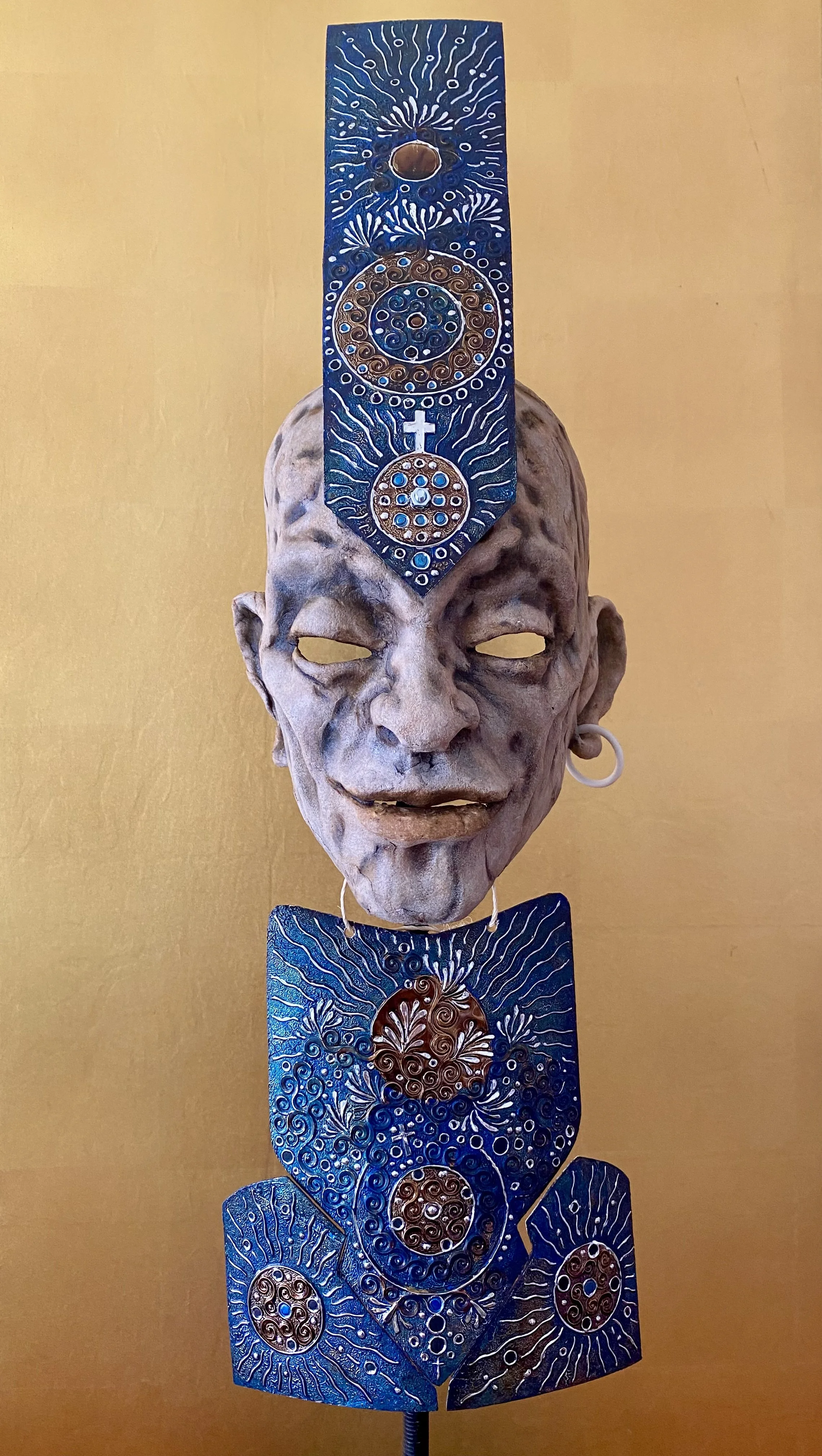 A wall sculpture featuring a detailed human face with closed eyes and an earring, topped and below by decorative blue and gold panels with intricate patterns and symbols.