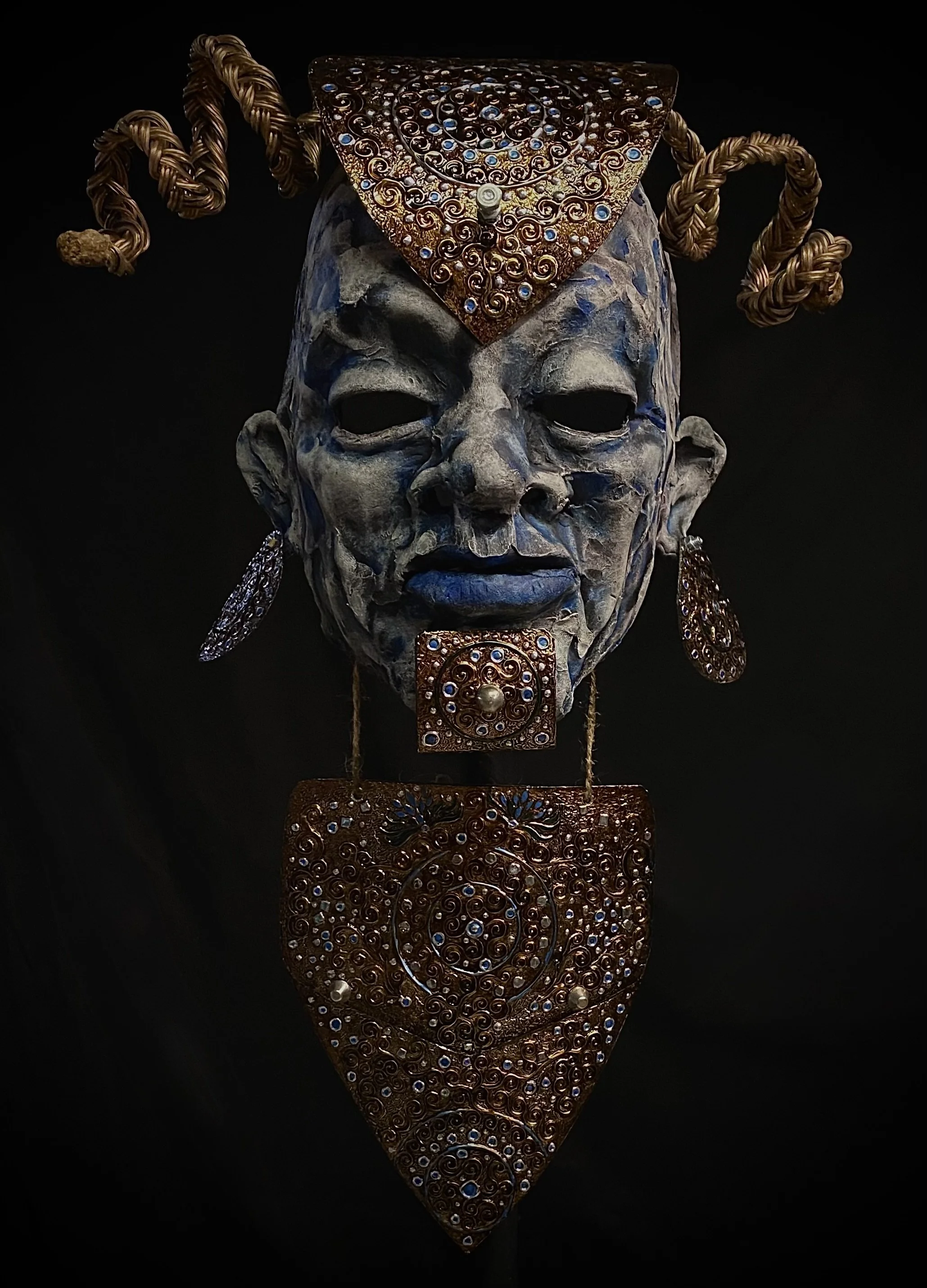 An artistic mask combining a human face with intricate jewelry adornments and a headdress, featuring metallic and gemstone details.