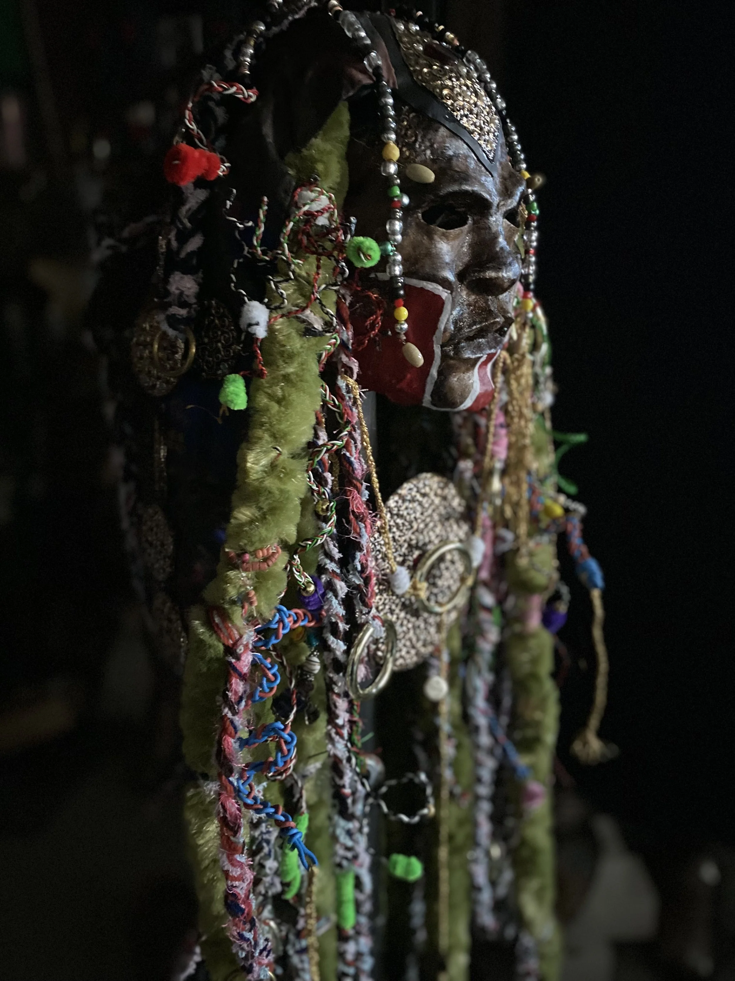 A traditional African ceremonial mask adorned with various colorful beads, fabric strips, and decorative embellishments, with a dark background.