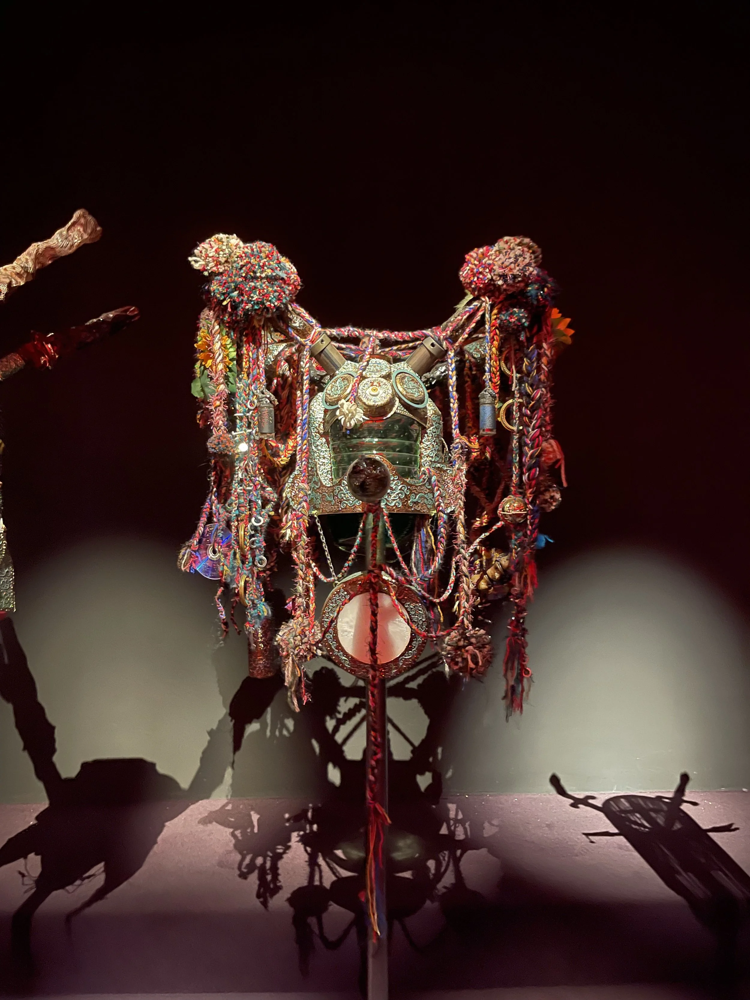 A decorative, ornate mask hanging on a dark wall, adorned with colorful beads, tassels, and fabric.