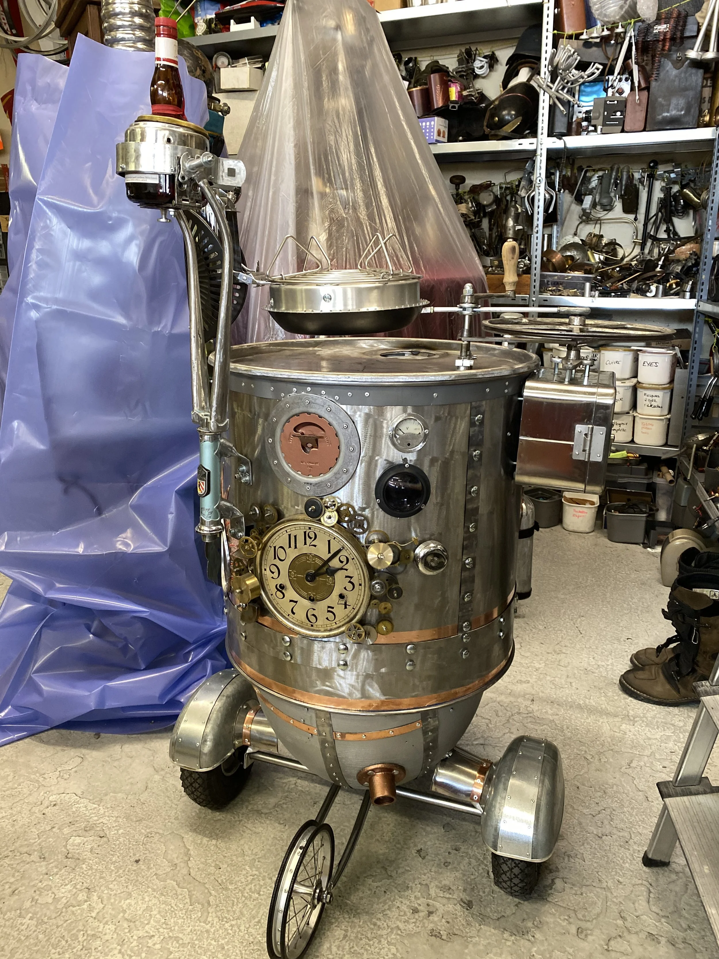 A vintage espresso machine with a clock, gauges, and knobs, made of metal with copper accents. It has wheels and is located in a workshop with shelves of tools and equipment in the background.