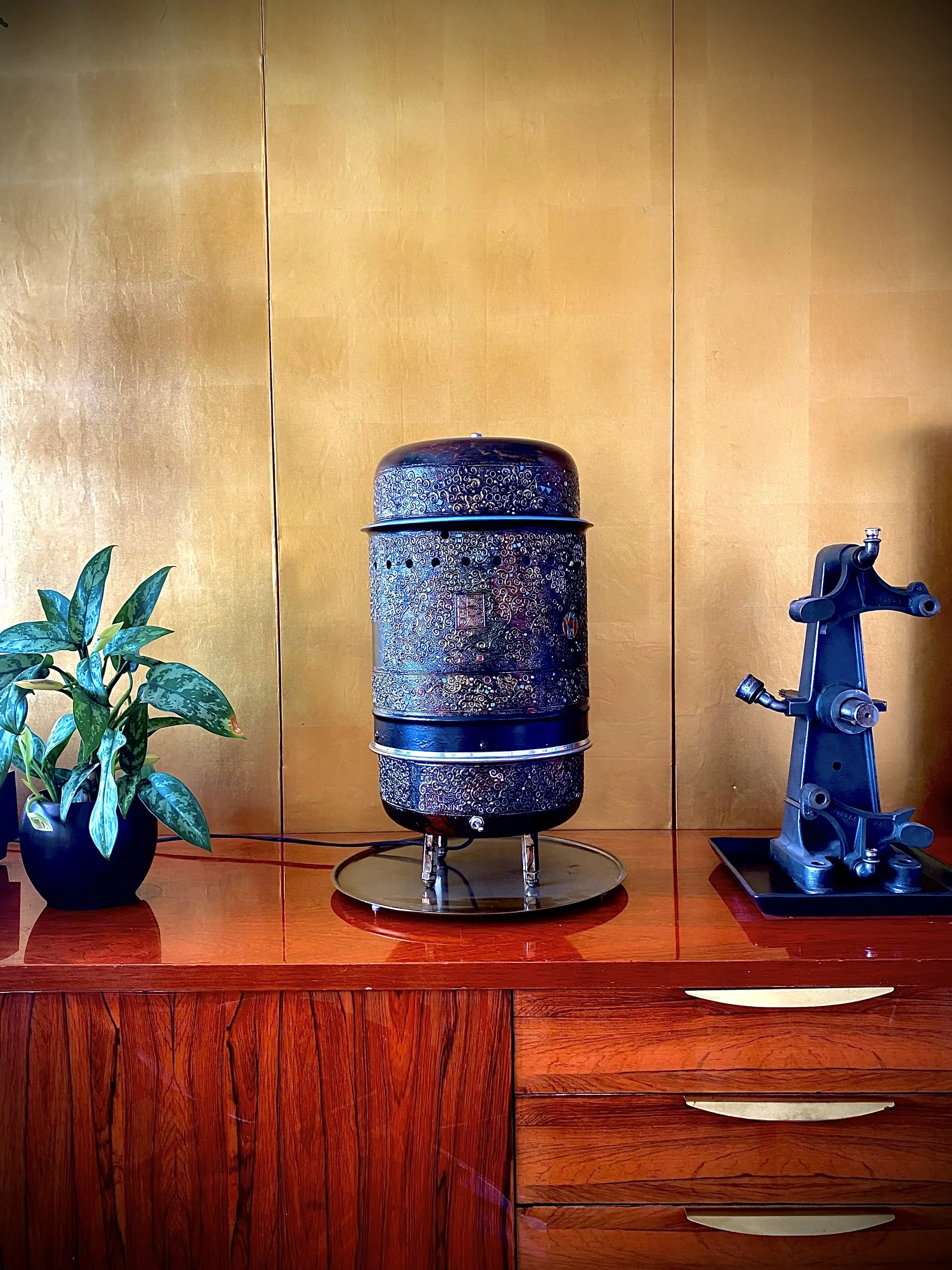 Decorative metallic container with intricate pattern, a potted green plant, and a vintage black microscope on a wooden cabinet with gold panels in the background.