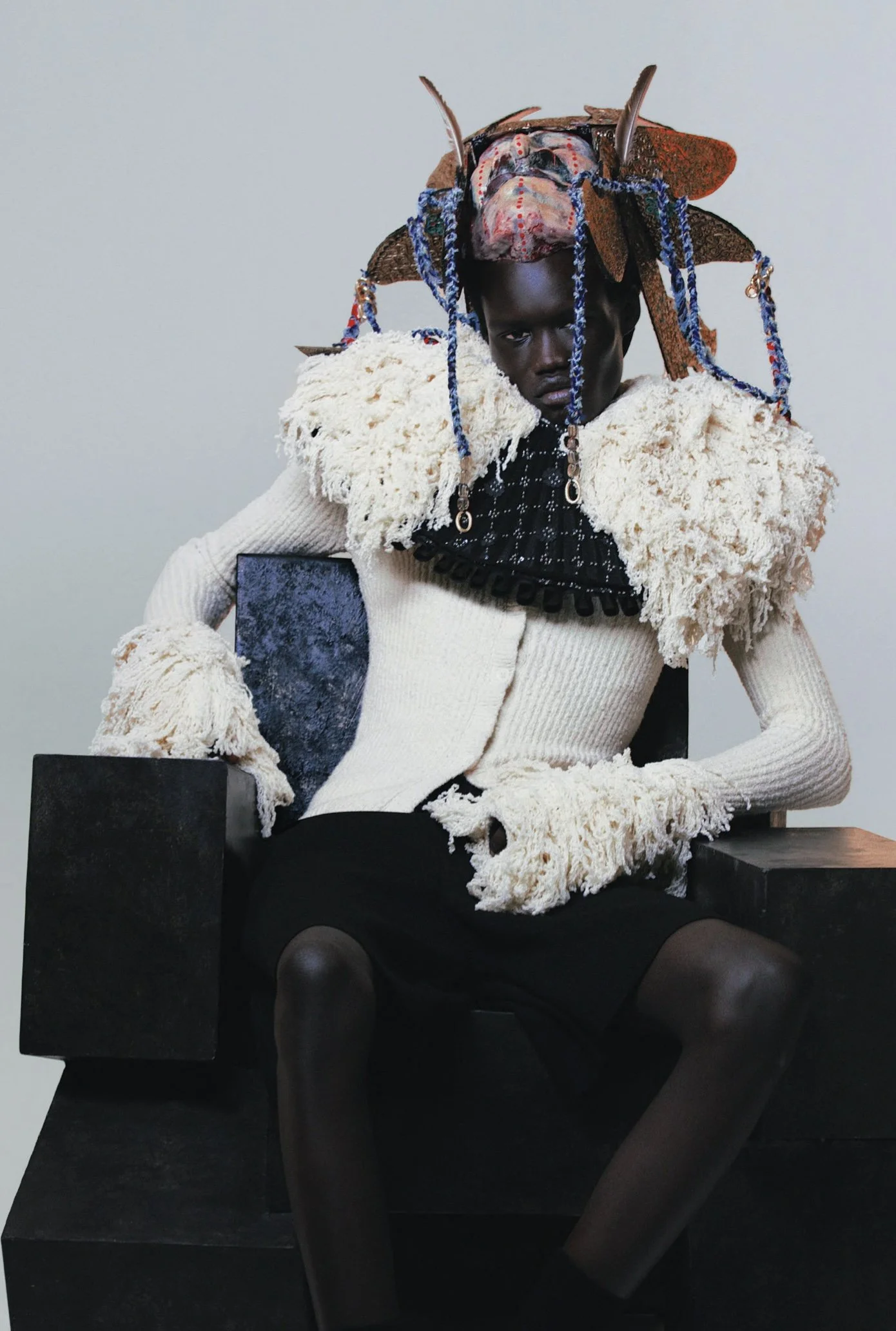 A dark-skinned person is sitting on a black chair, wearing a designer outfit with textured white and black fabrics, extravagant accessories, and a headpiece with feathers and beads, against a plain light-colored background.