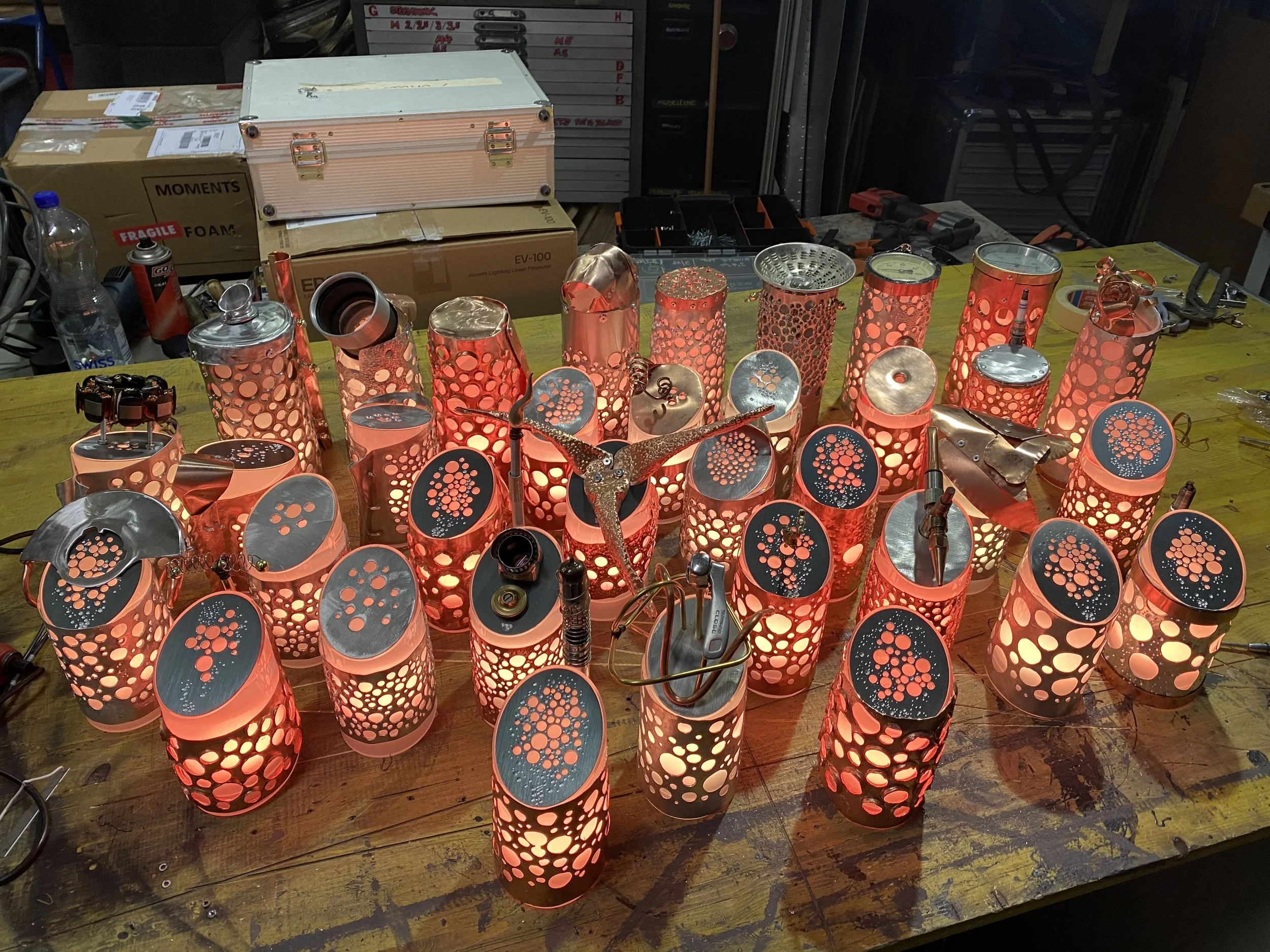 A wooden workbench covered with assembled decorative table lamps made from metal cans with circular cutouts, lit from within. Various tools and boxes are in the background.