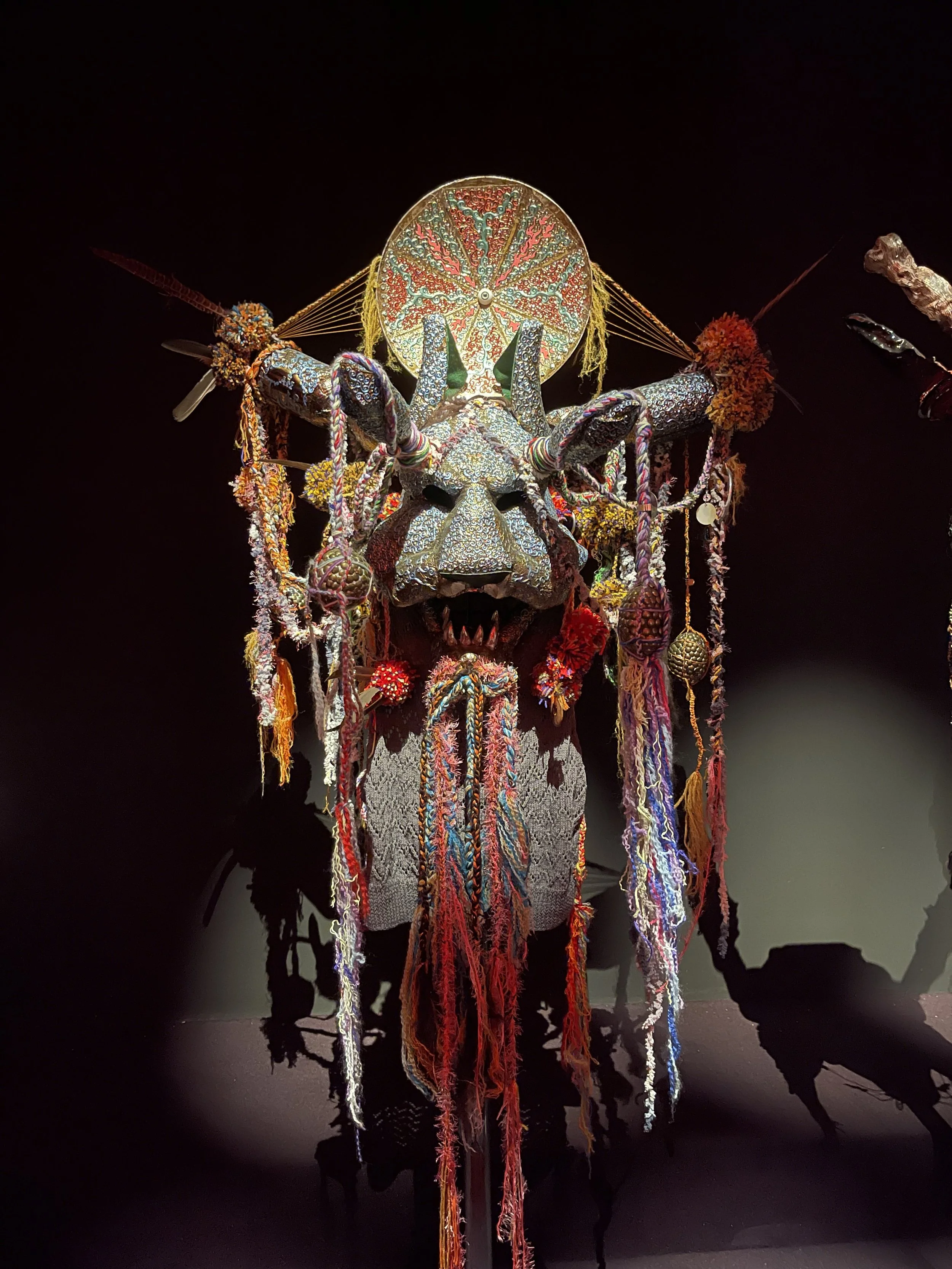 An elaborate, colorful, tribal-style mask with a fierce animal face, decorated with yarn, beads, and ornaments, displayed against a black background.