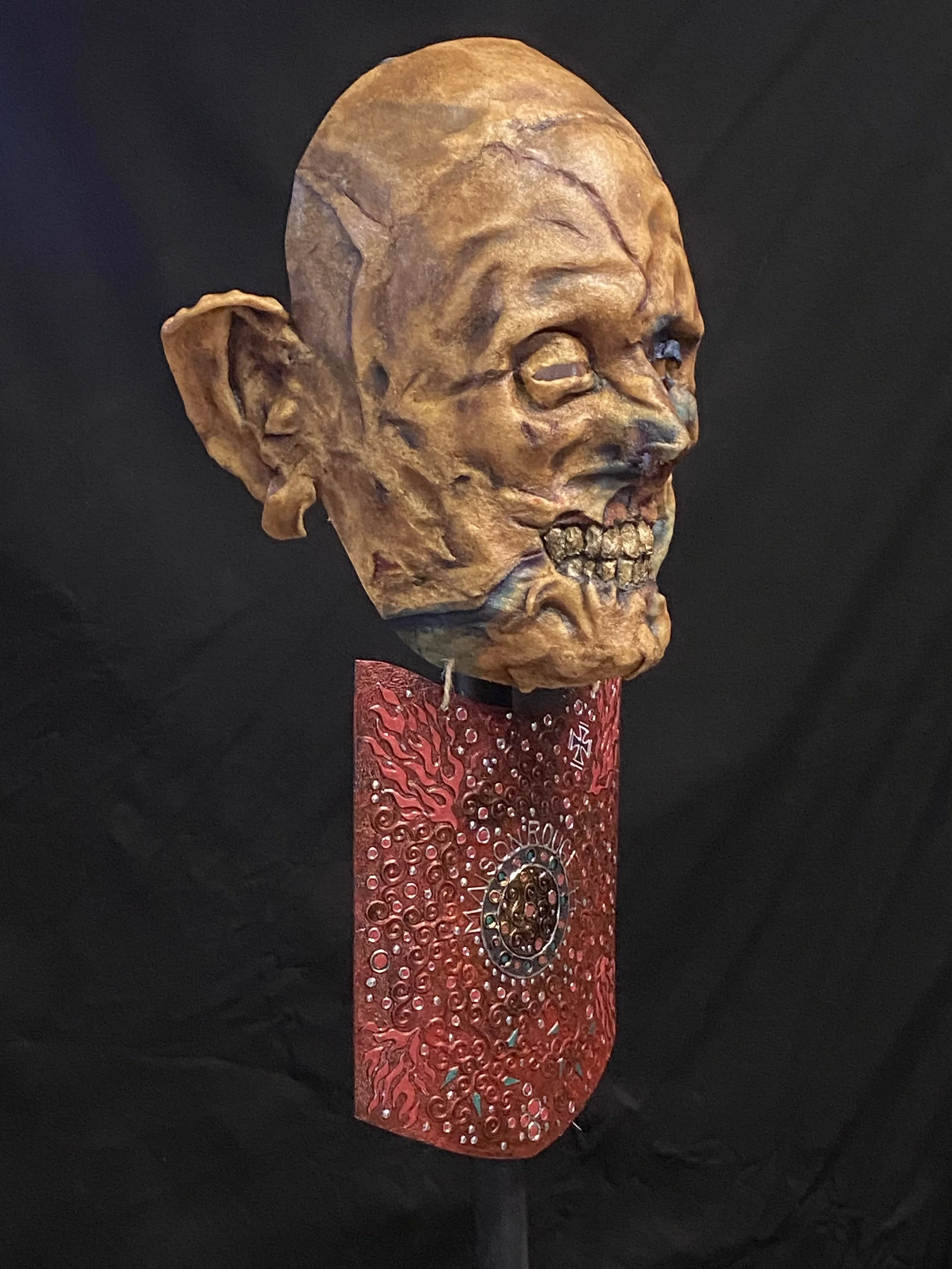 A sculpture of a human skull with exaggerated features, mounted on a red, decorative plaque against a black background.