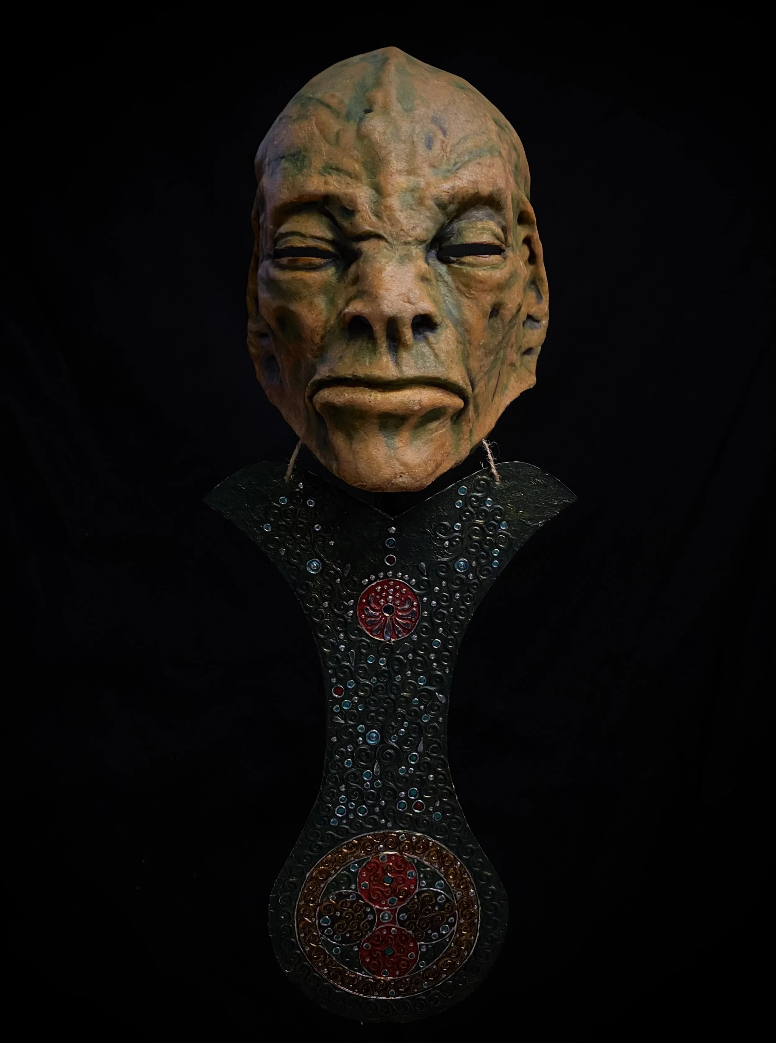 A carved mask resembling a human face with closed eyes and textured skin, mounted on a decorative stand with intricate patterns and red, blue, and gold embellishments.