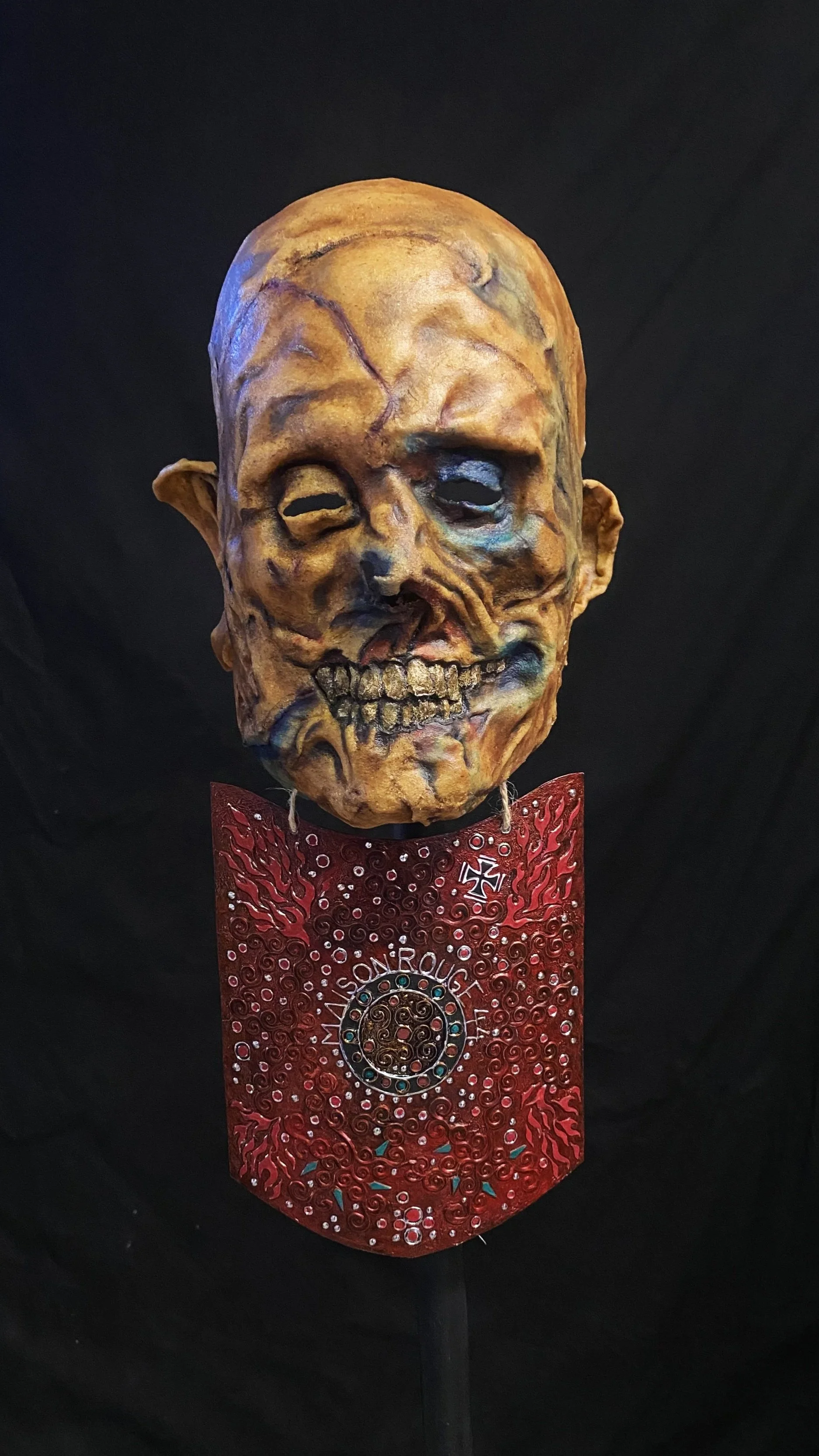 A Halloween mask resembling a decayed skull with hollow eyes and missing teeth, mounted above a red decorative shield with intricate patterns and the words "Maison Rouge".