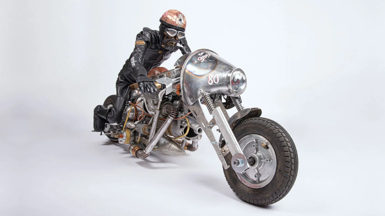 A person in black leather gear and helmet is riding a vintage-style motorcycle with a metallic body and large front wheel