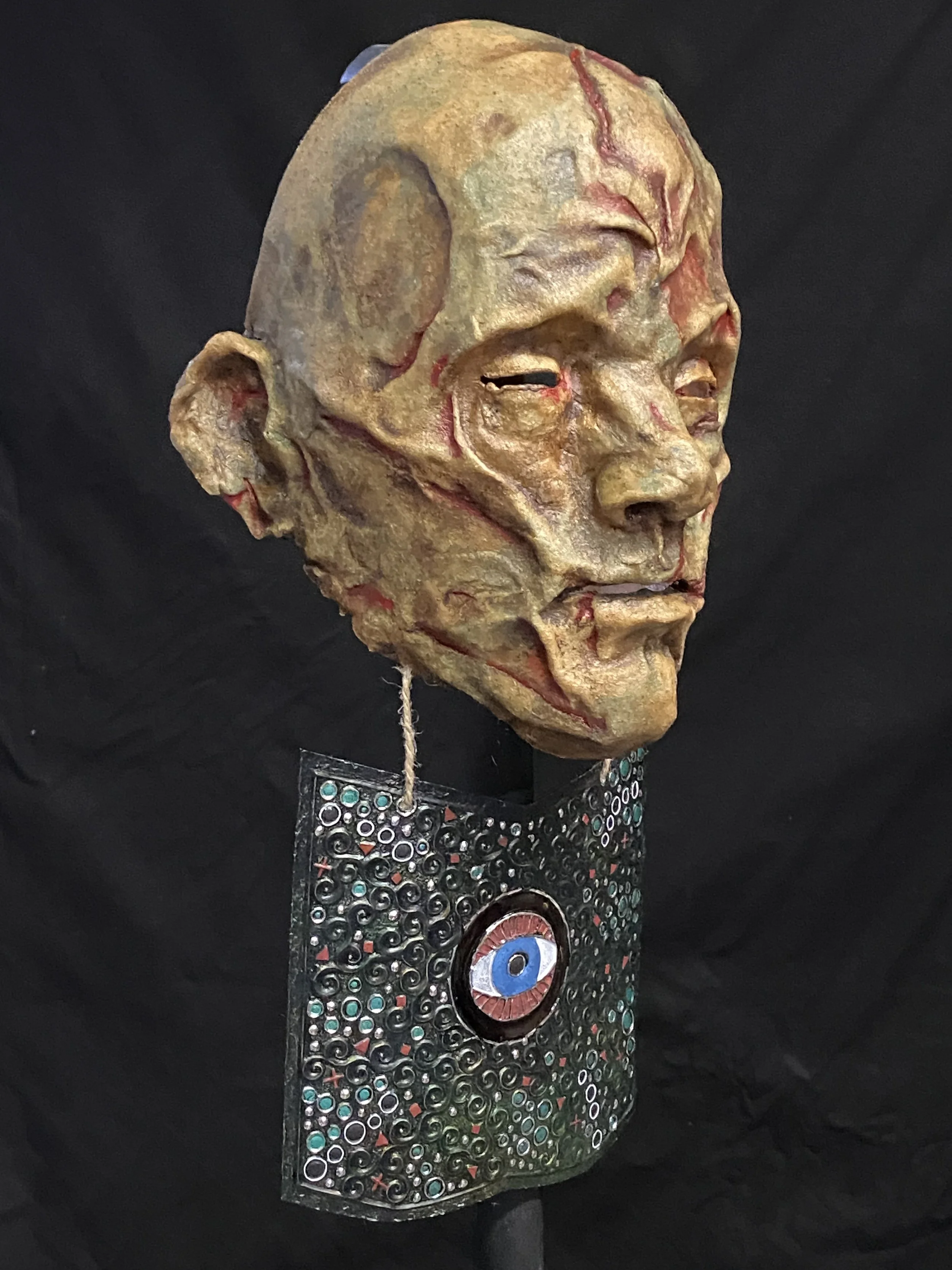 An abstract sculpture of a human face with a rough, textured surface, mounted on a stand with decorative elements and a central eye motif.