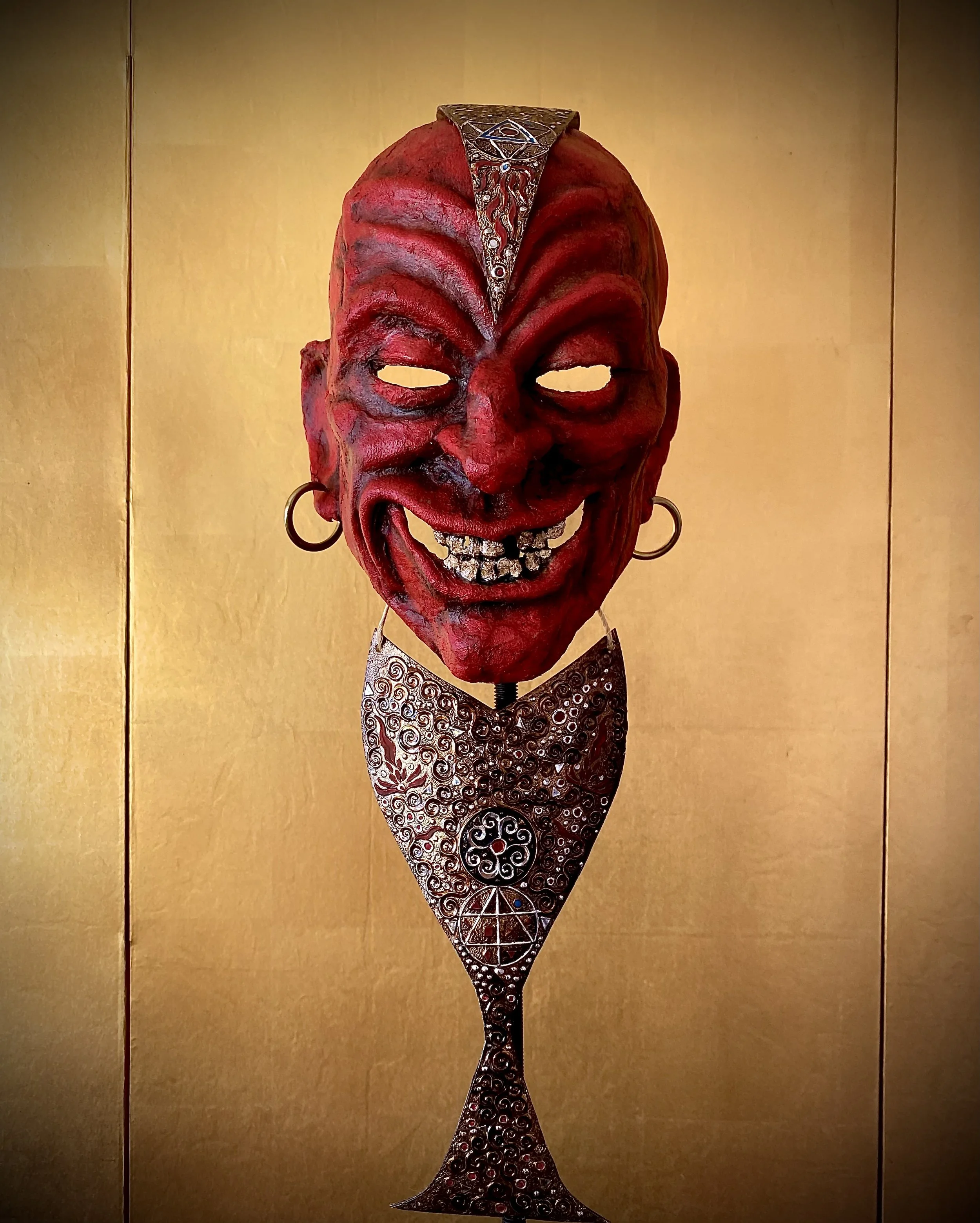 A red, leather horned mask with exaggerated facial features and a wide, toothy grin, dressed with large hoop earrings and a decorative metal necklace, displayed against a gold-colored background.