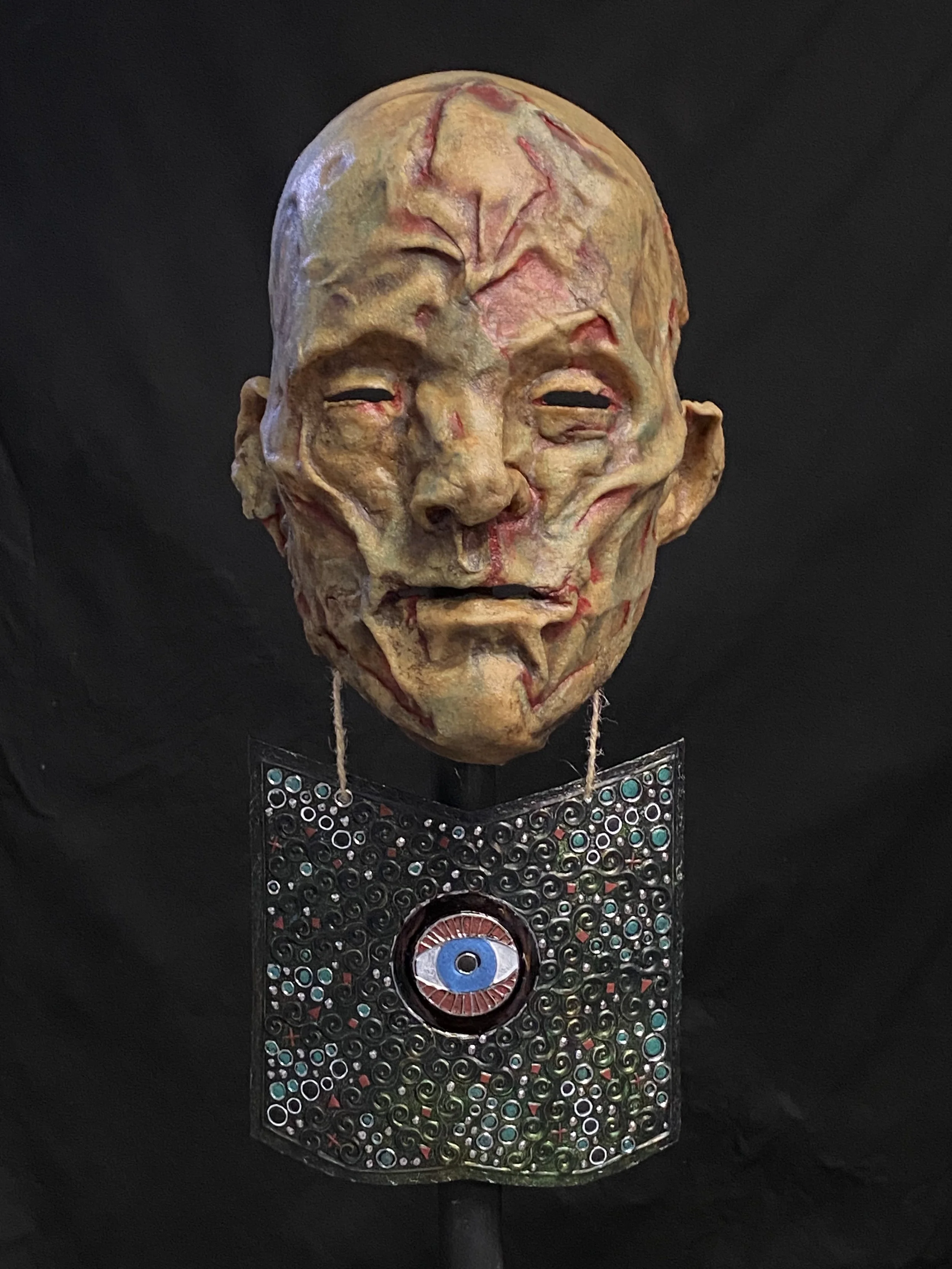 Artistic mask resembling a wrinkled, aging face with closed eyes and an open mouth, hanging above a decorative metal panel with a large central eye design.